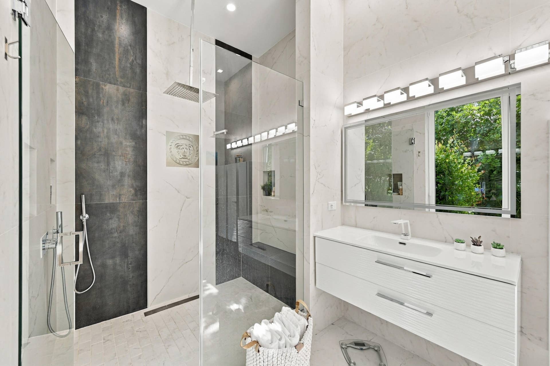 110 NE 16TH Place, Fort Lauderdale, FL 33305 Glass-enclosed shower with a rainfall showerhead and handheld sprayer, contrasting dark-toned shower wall, floating vanity with an integrated sink, and extensive wall-mounted mirror with overhead lighting