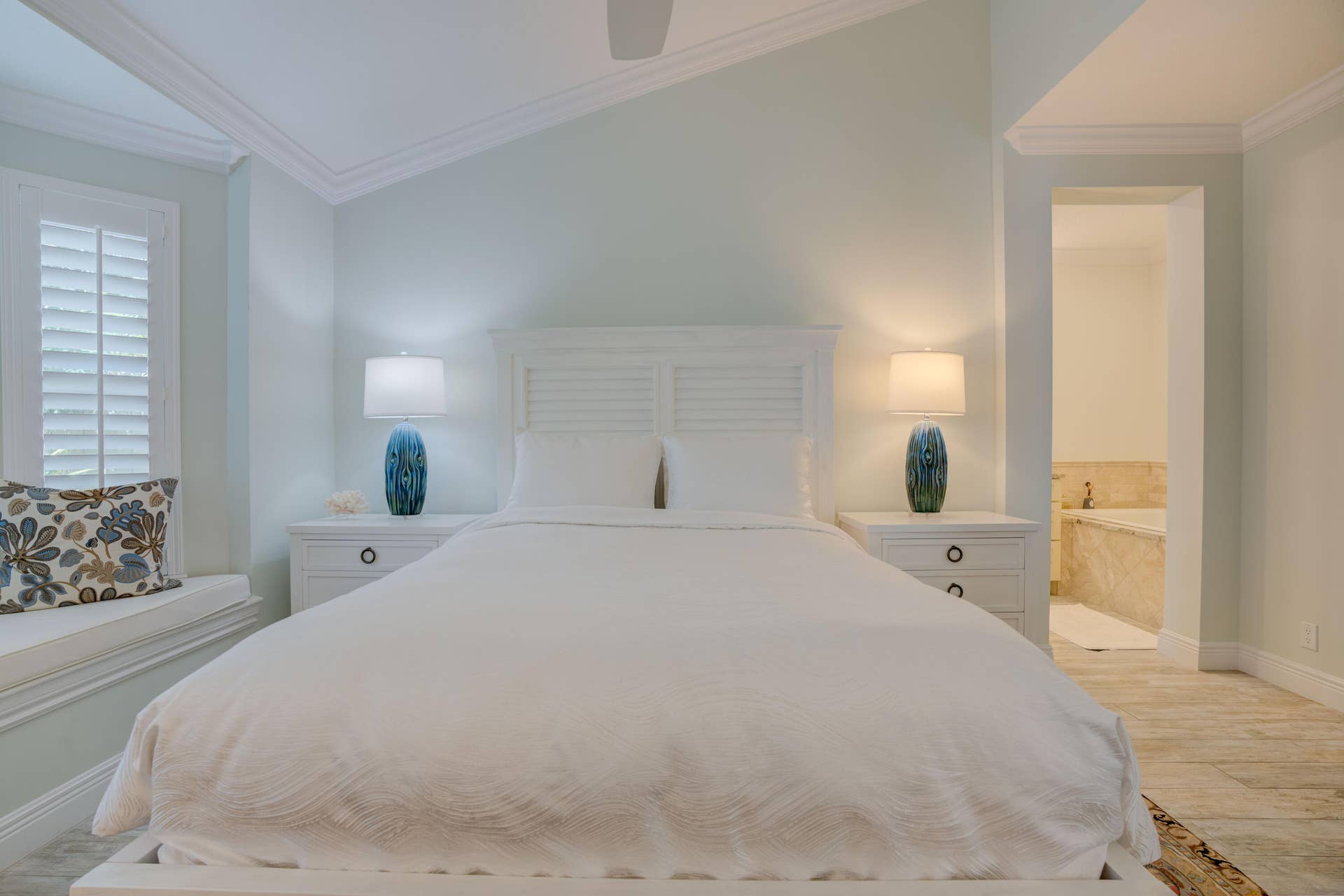 2043 Staysail Lane, Jupiter, Jupiter, FL 33477 Tray ceiling with crown molding, window seat with plantation shutters, louvered headboard, white bedside tables, and an adjacent bathroom with a built-in tub