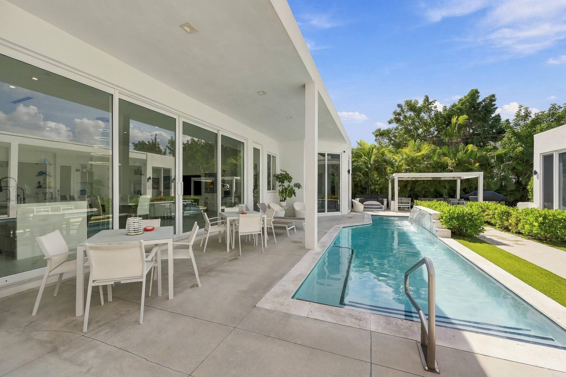 Expansive outdoor living area featuring a covered patio, a rectangular swimming pool with integrated steps, a white pergola, and manicured landscaping