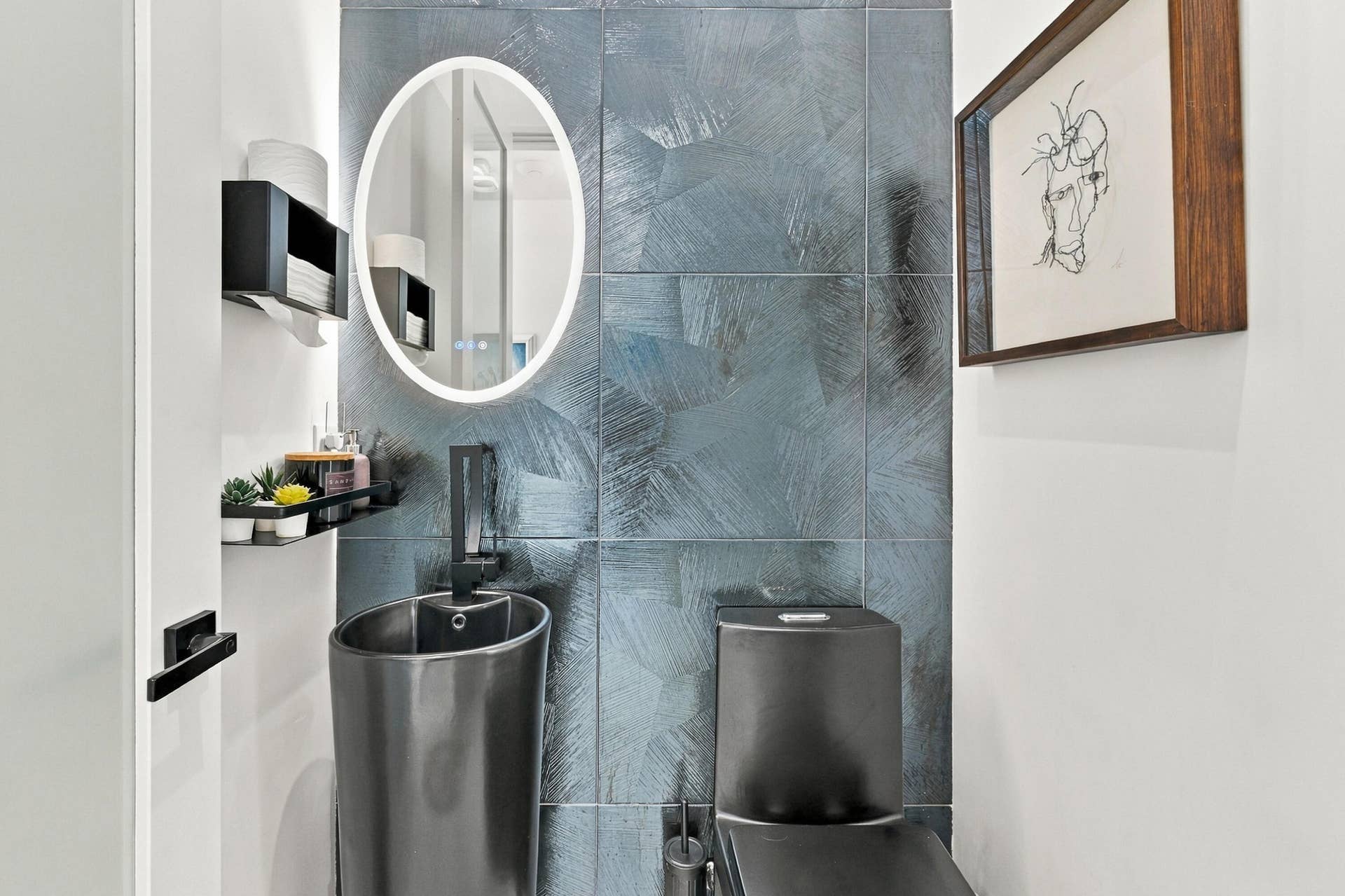 Textured wall tiling, pedestal sink, illuminated oval mirror, matte black fixtures, and a black toilet