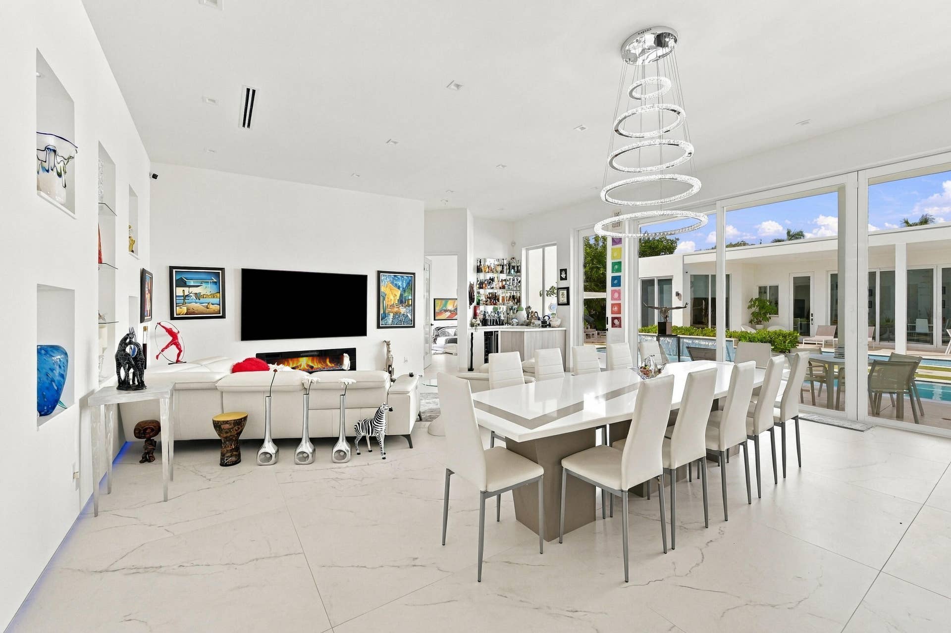 Expansive open-concept living area featuring white tile flooring, a linear electric fireplace, built-in wall niches, a multi-tiered chandelier, and large sliding glass doors