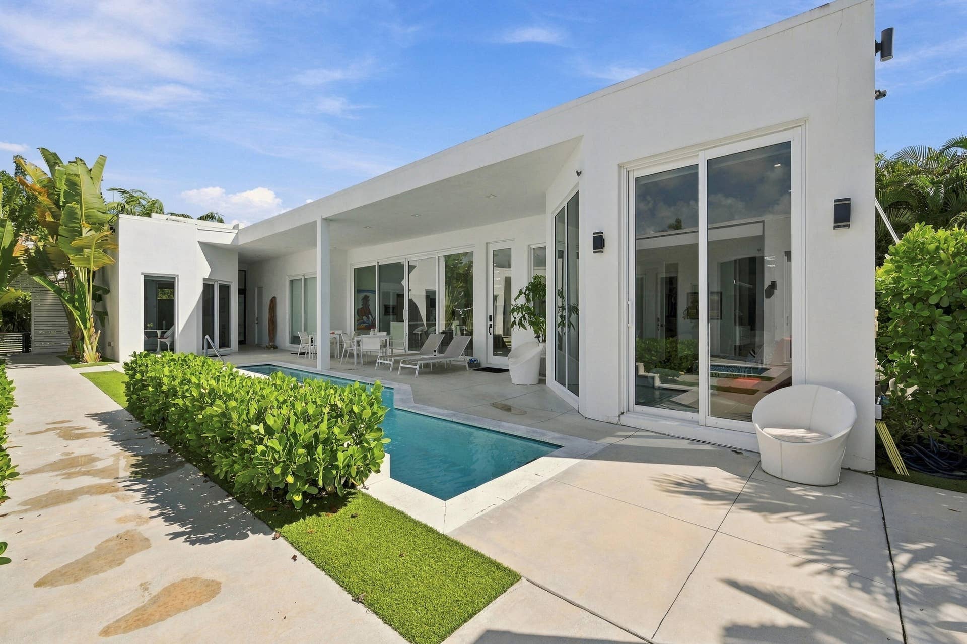 Modern exterior featuring a rectangular swimming pool, concrete patio, white stucco facade, multiple sliding glass doors, and manicured landscaping