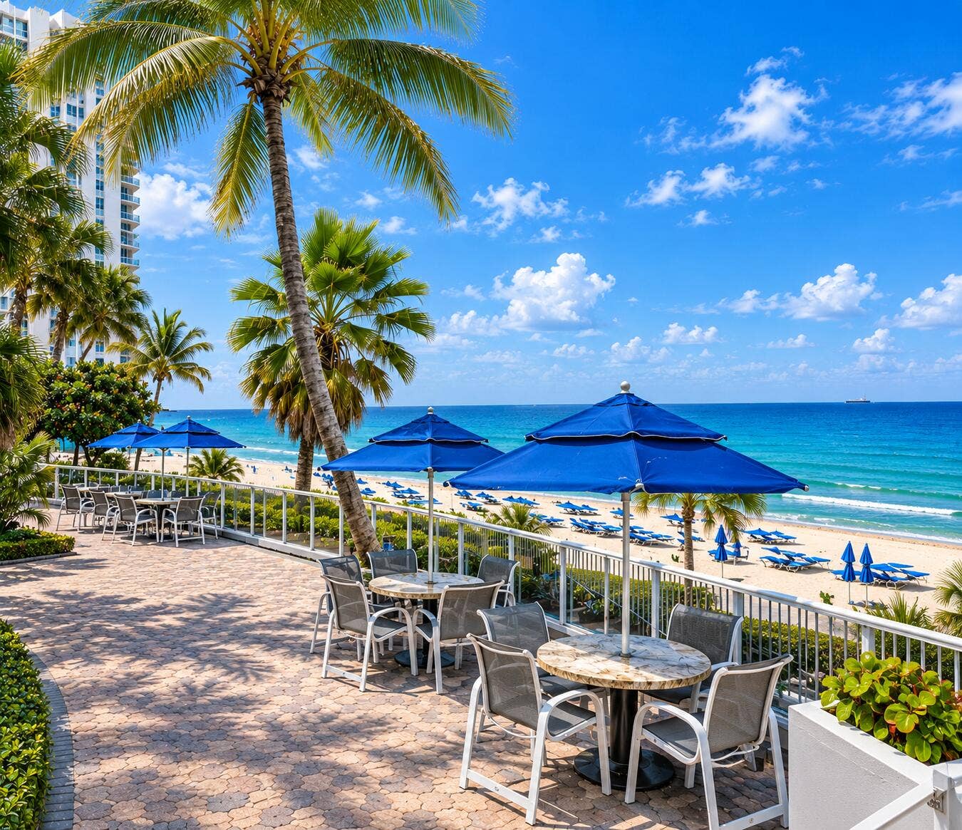 Paved patio area with multiple tables and chairs, surrounded by lush landscaping and palm trees, with a beach and ocean backdrop