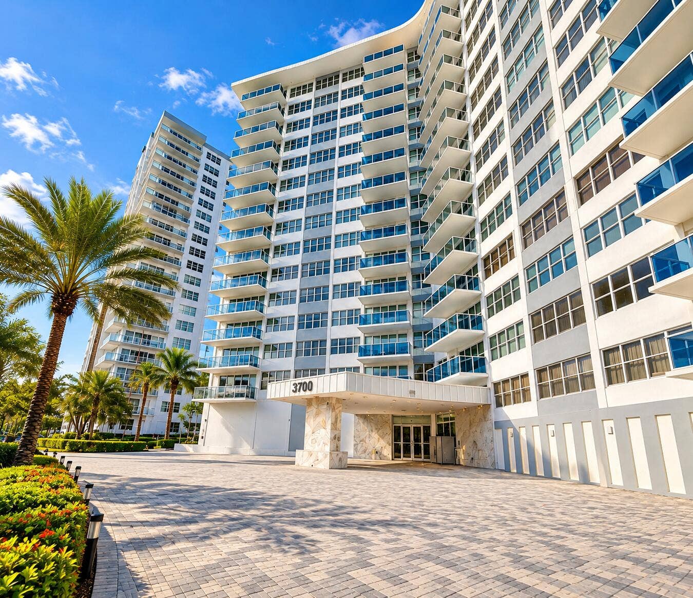 Modern high-rise building featuring expansive balconies with clear glass railings, a stone-clad entrance, and a paver driveway