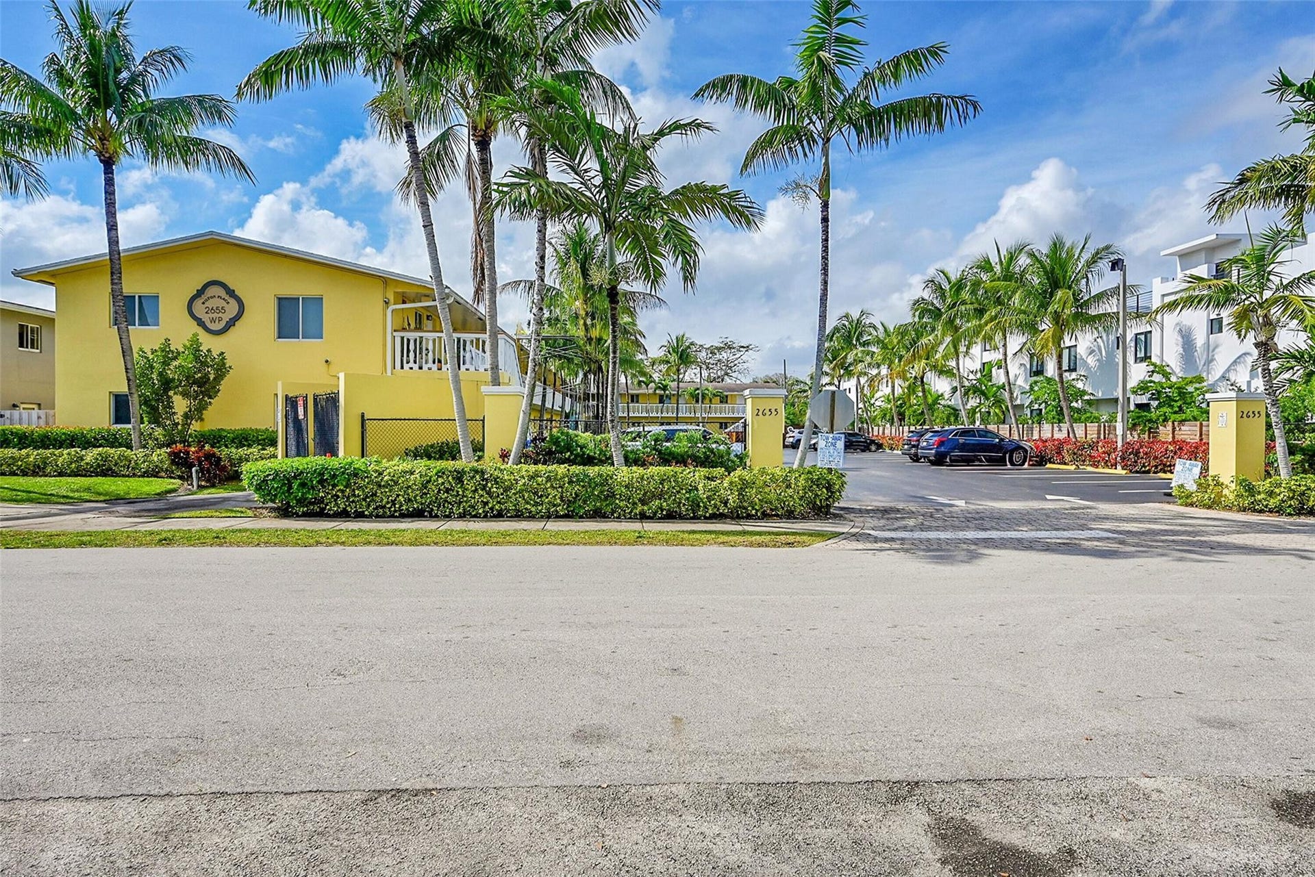 2655 NE 8th Avenue, #Apt 210, Wilton Manors, FL 33334 Yellow stucco exterior, white balcony railing, mature palm trees, lush landscaping, and prominent signage