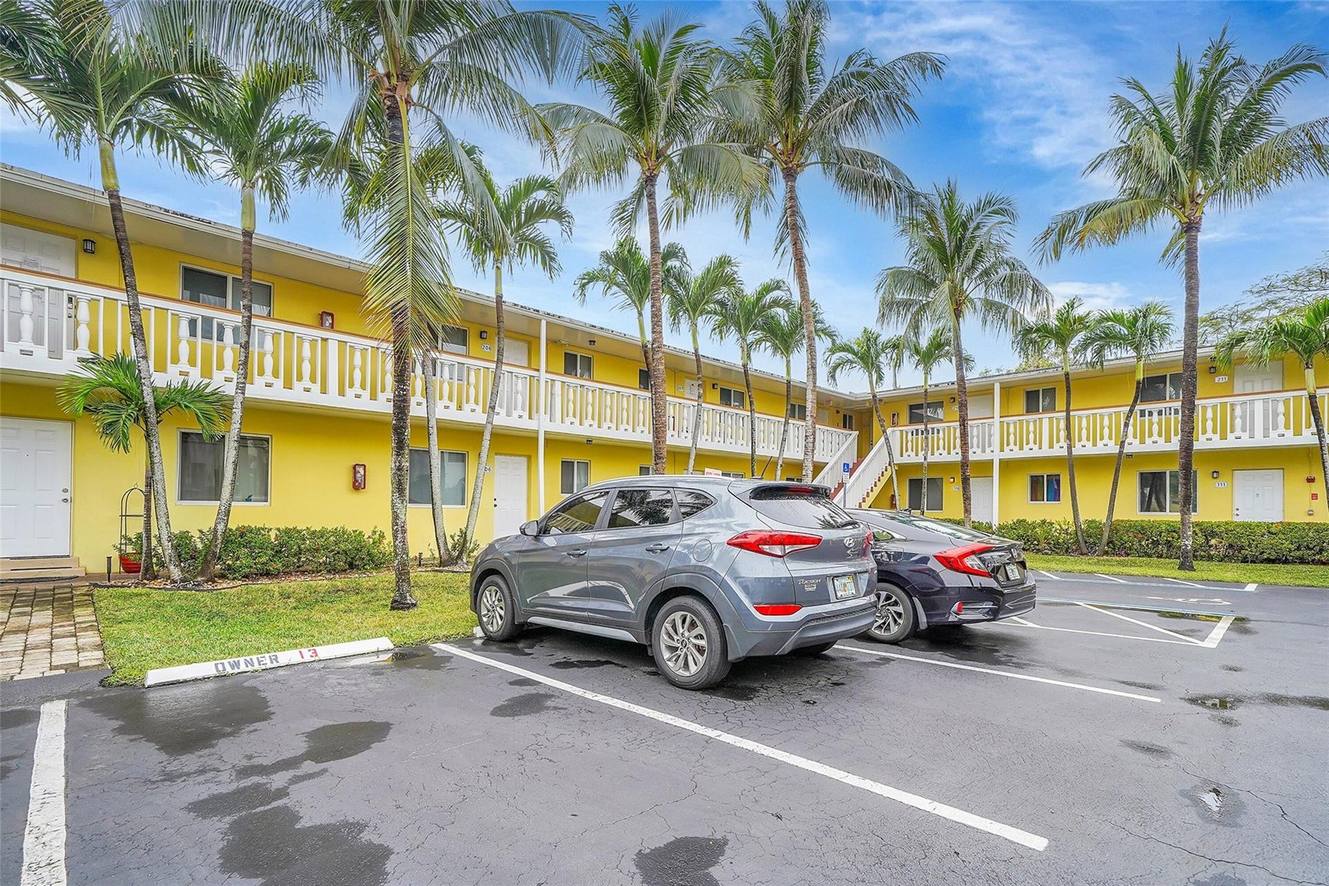 2655 NE 8th Avenue, #Apt 210, Wilton Manors, FL 33334 Exterior showcasing a yellow facade, white railings, mature palm trees, and an asphalt parking area with marked spaces