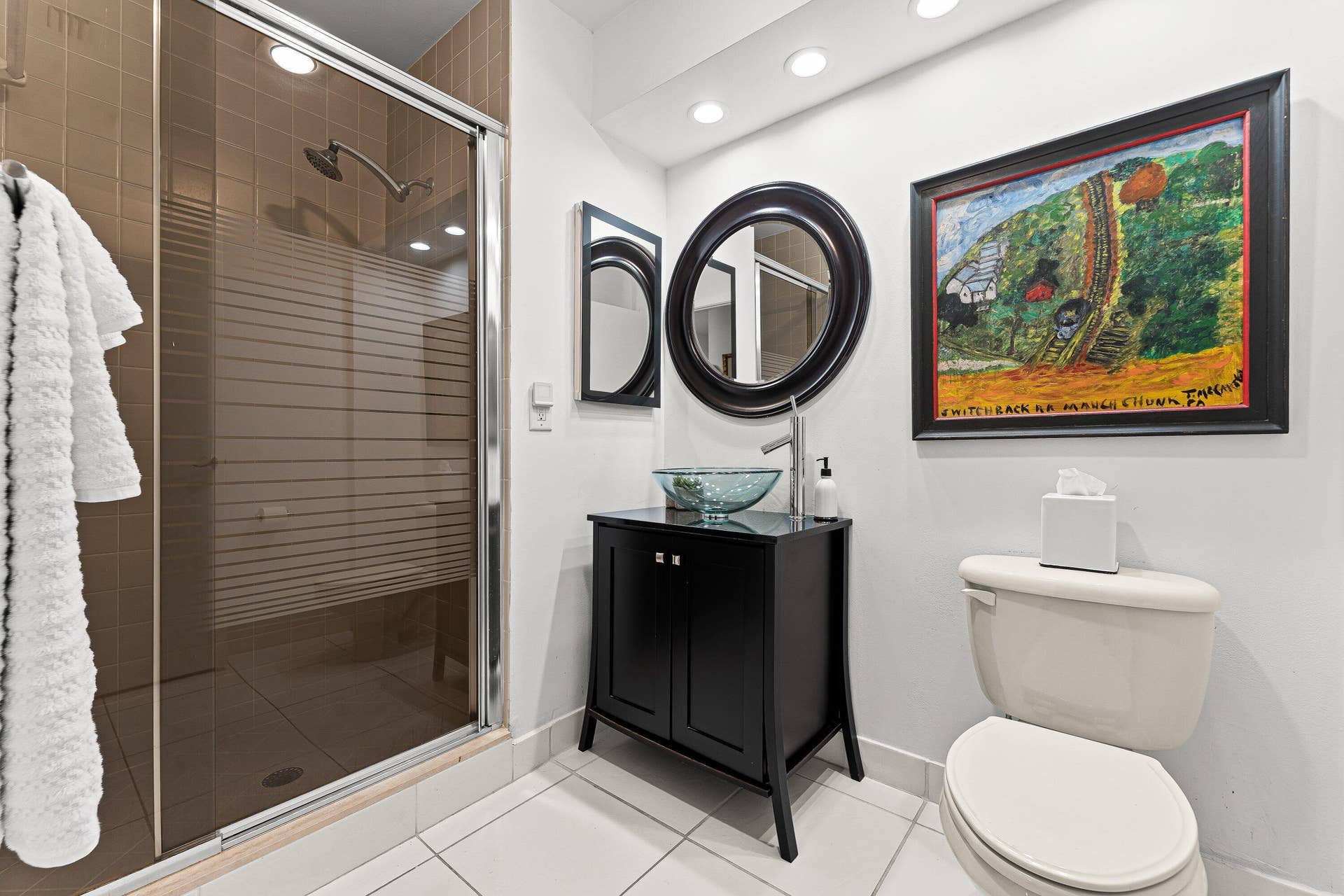 Walk-in shower with a glass enclosure and tiled walls, black vanity with a vessel sink, round wall-mounted mirror, and recessed lighting