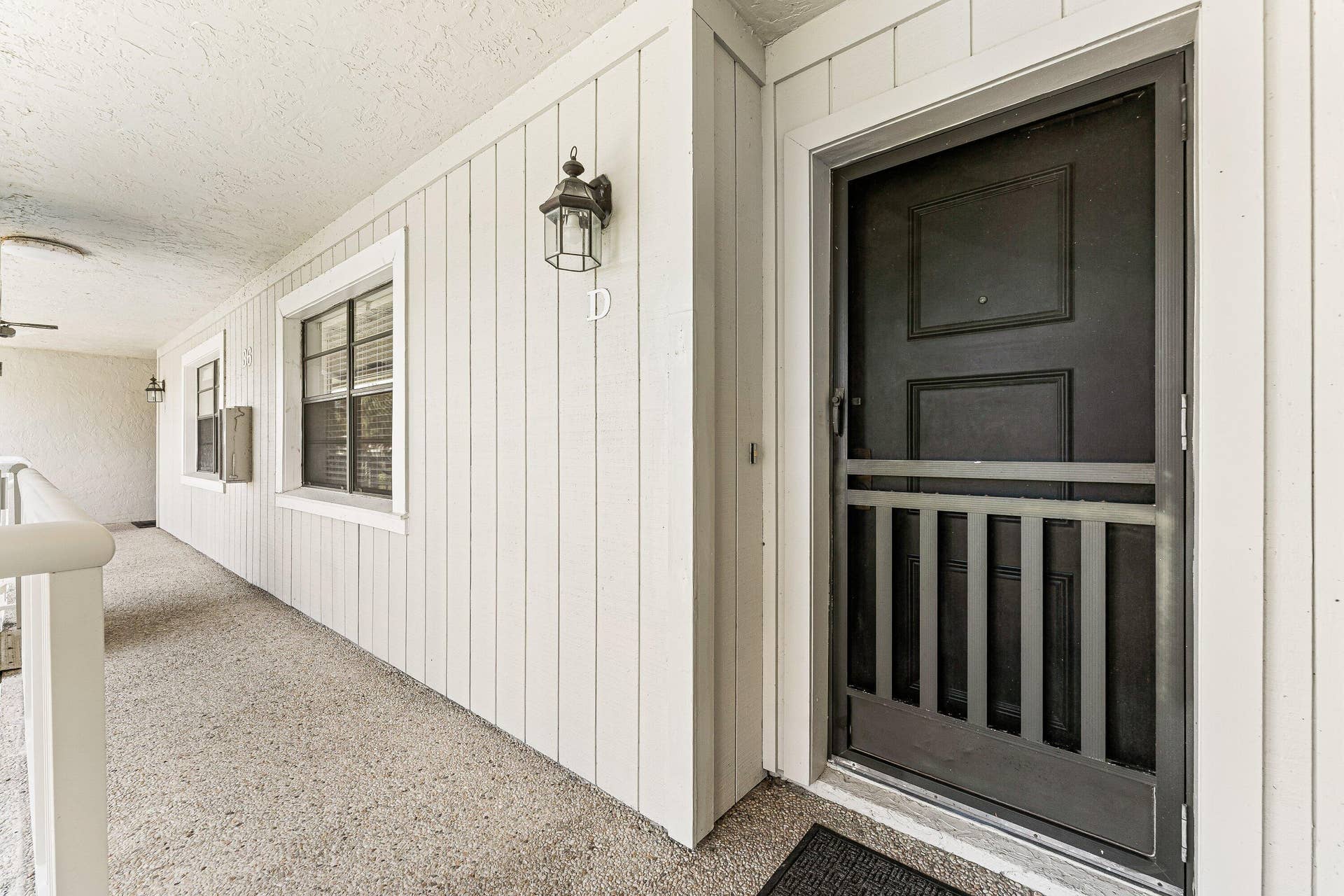 Covered exterior walkway featuring a textured floor, vertical siding, decorative wall sconce, and a security screen door
