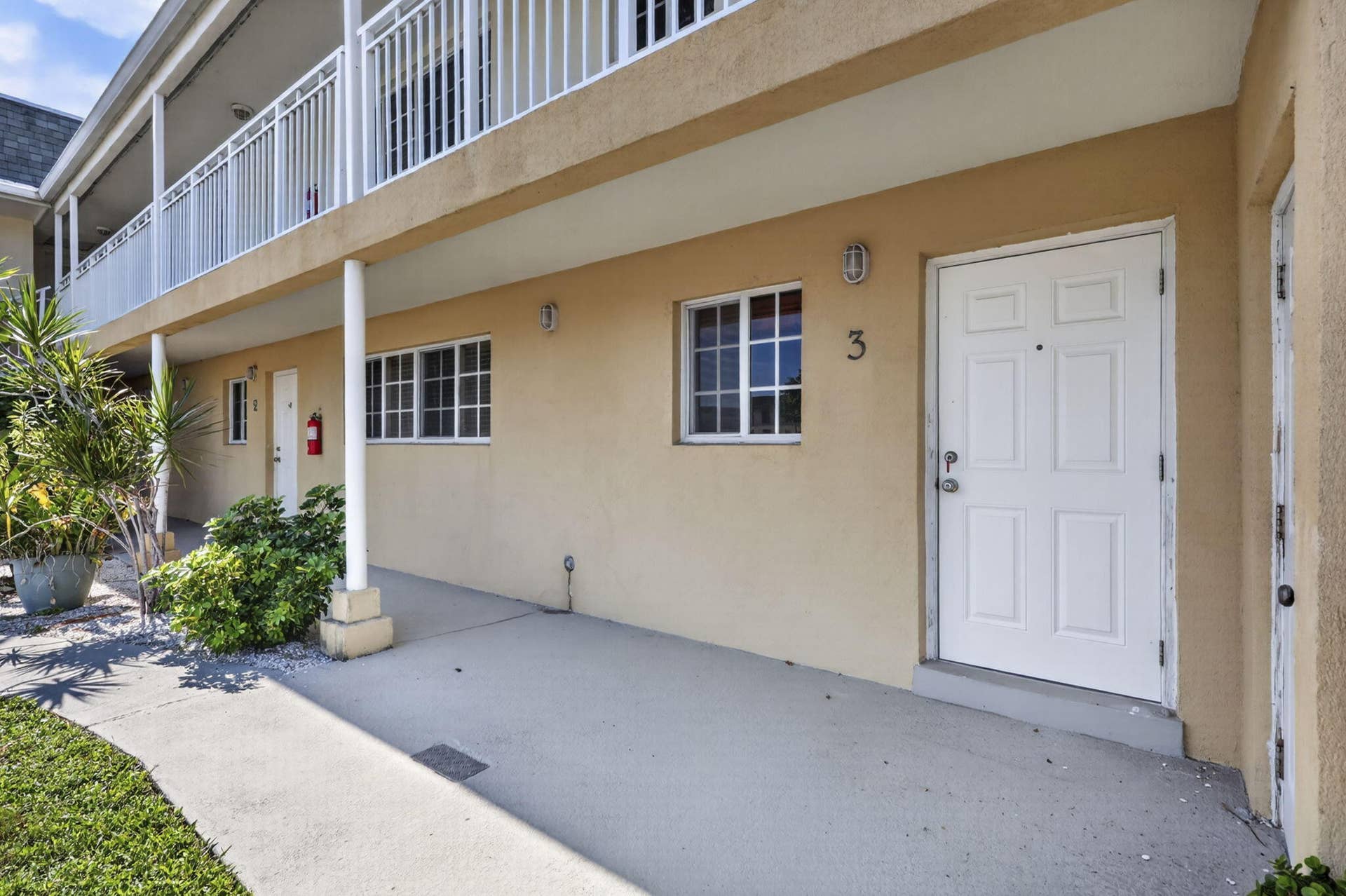 405 SOUTHWIND Drive, #3, North Palm Beach, FL 33408 Exterior facade featuring a smooth stucco finish, covered concrete walkway, second-level balcony with white railings, grid-patterned windows, and wall-mounted exterior lighting