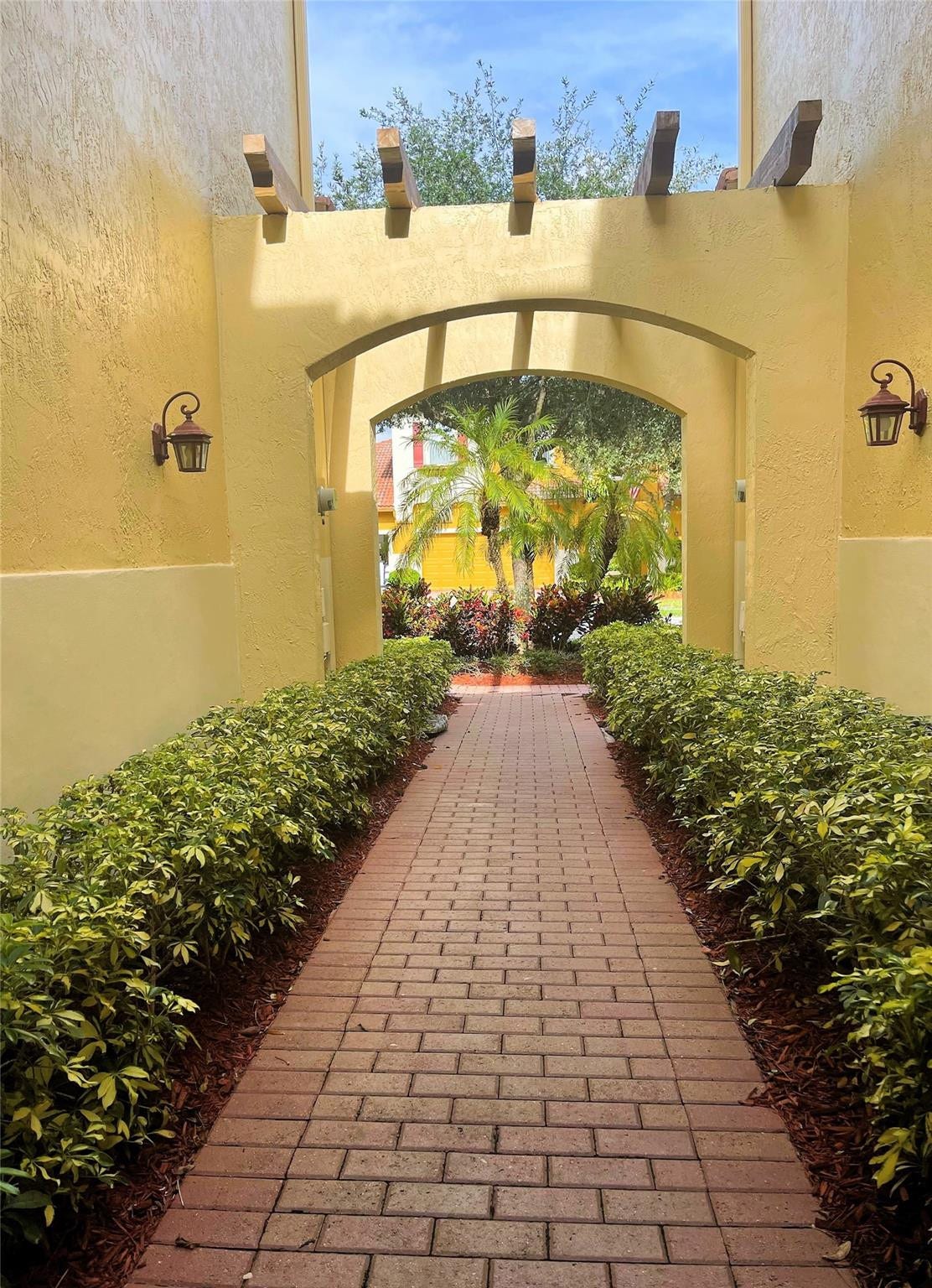 5780 NW 120TH Avenue, Coral Springs, Coral Springs, FL 33076 Brick paver walkway lined with manicured hedges, featuring a stucco archway, decorative wood-beam accents, and wall-mounted exterior sconces