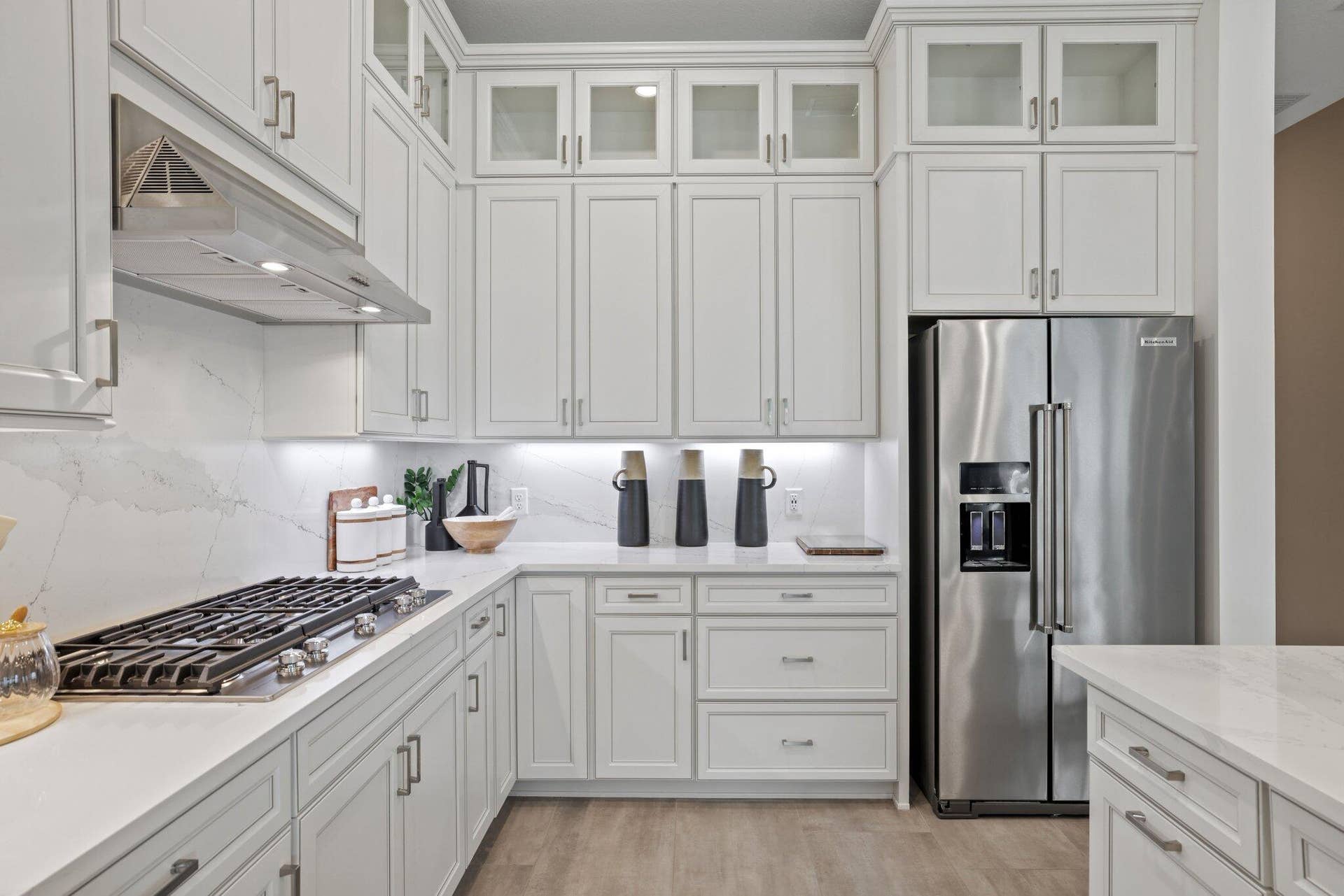 205 GEILLIS Path, Delray Beach, FL 33445 White shaker-style cabinetry with glass-front uppers, light-toned stone-finish countertops, full-height stone-finish backsplash, stainless steel gas cooktop with matching range hood, and integrated stainless steel refrigerator
