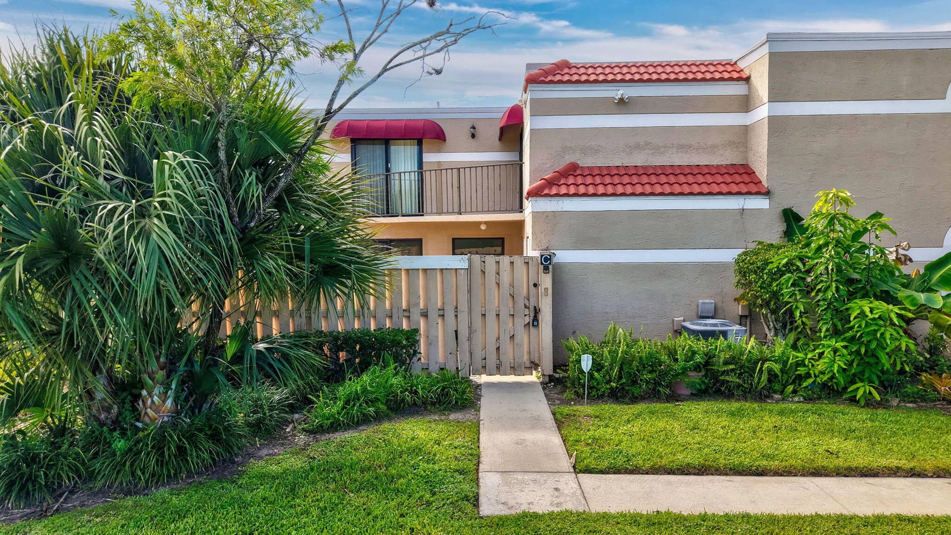 Stucco exterior with red tile roof accents, ground-level privacy fence, mature landscaping, second-story balcony with awning, and concrete walkway