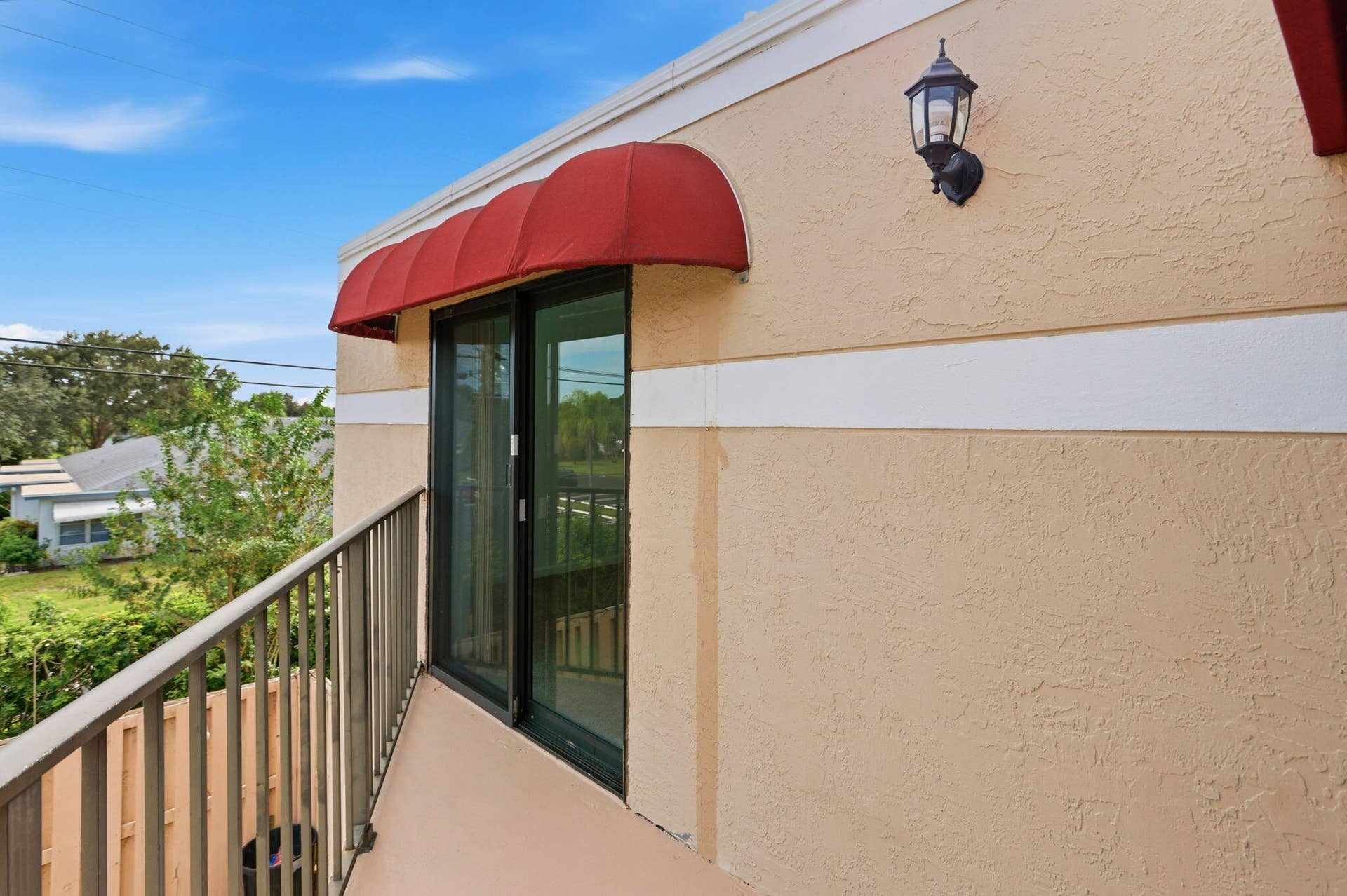 Balcony with metal railing, sliding glass door, red fabric awning, and exterior wall sconce