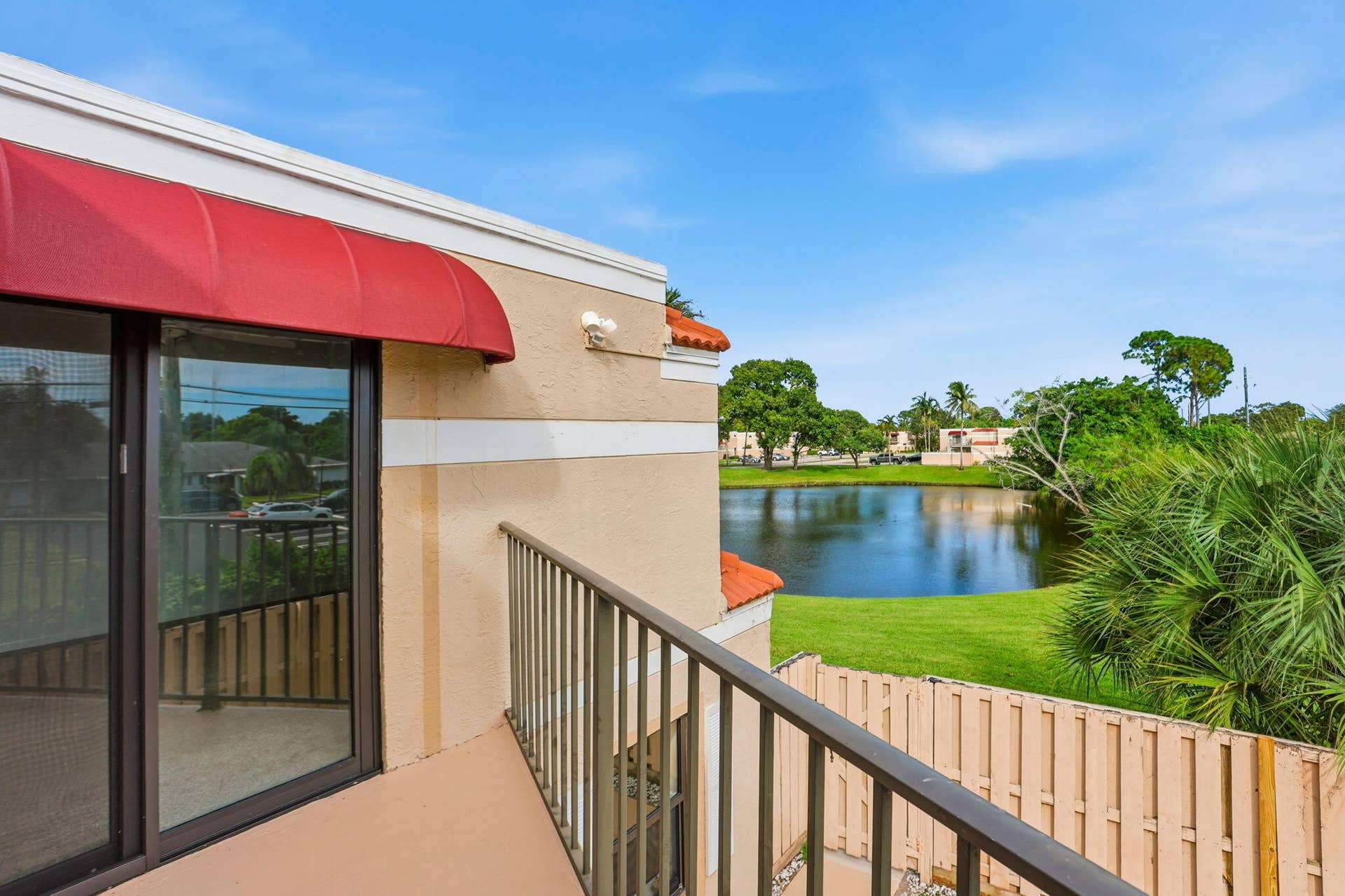 Balcony with metal railings, red awning, and sliding glass doors overlooking a body of water with lush landscaping