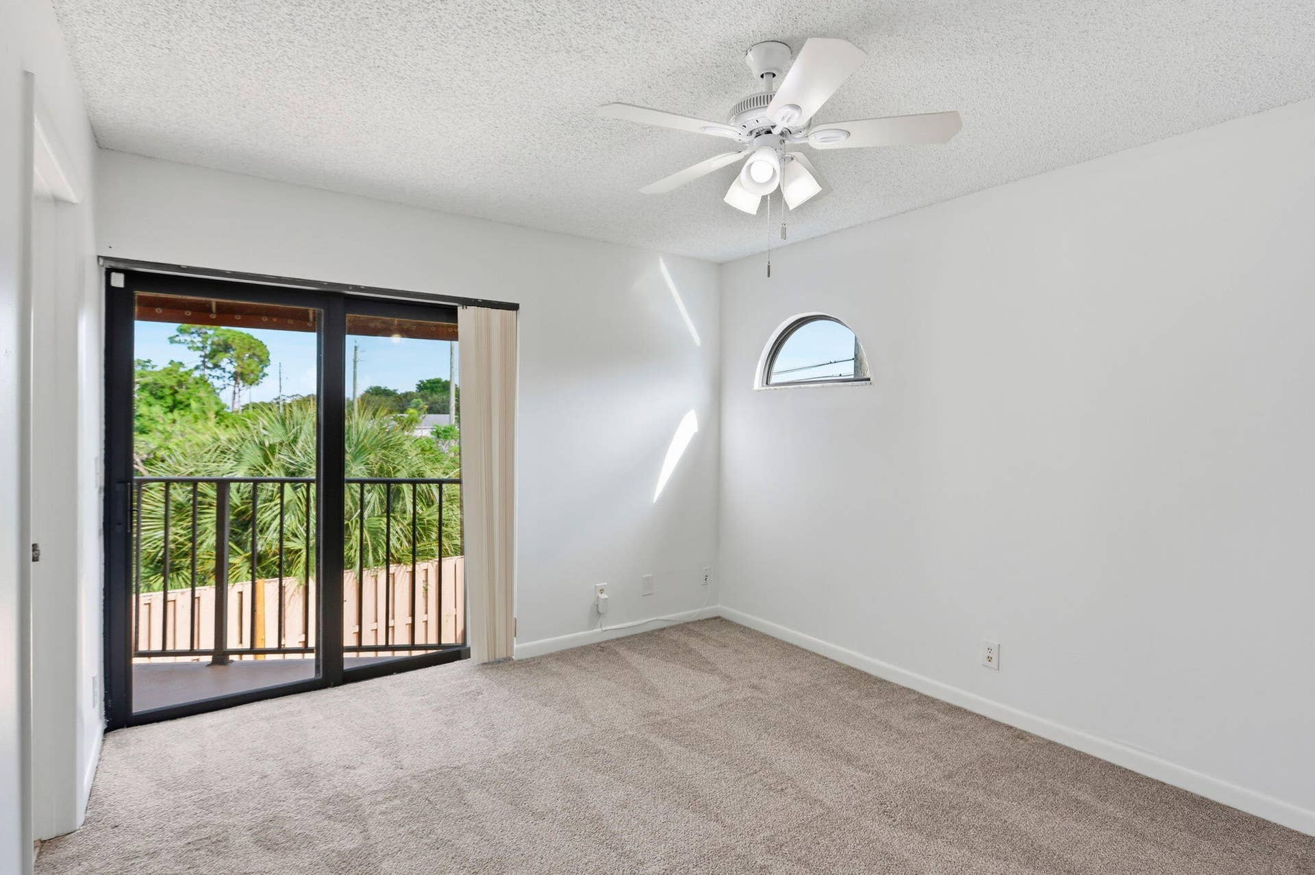 Carpeted interior with a ceiling fan, arched window, and sliding glass door leading to a balcony with black metal railings