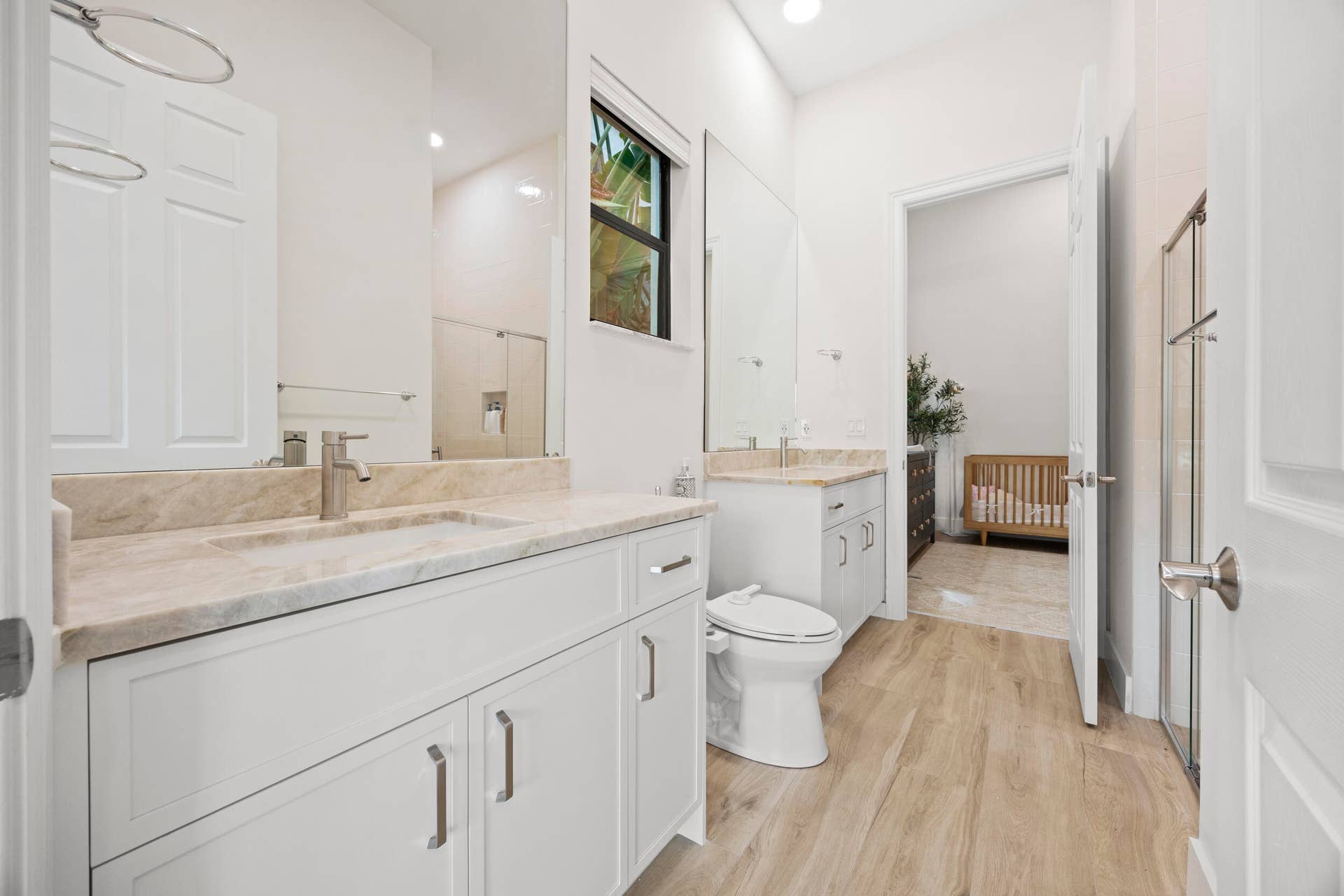 Dual-vanity bathroom featuring white shaker-style cabinetry, neutral stone countertops, wood-finish flooring, and expansive wall-to-wall mirrors