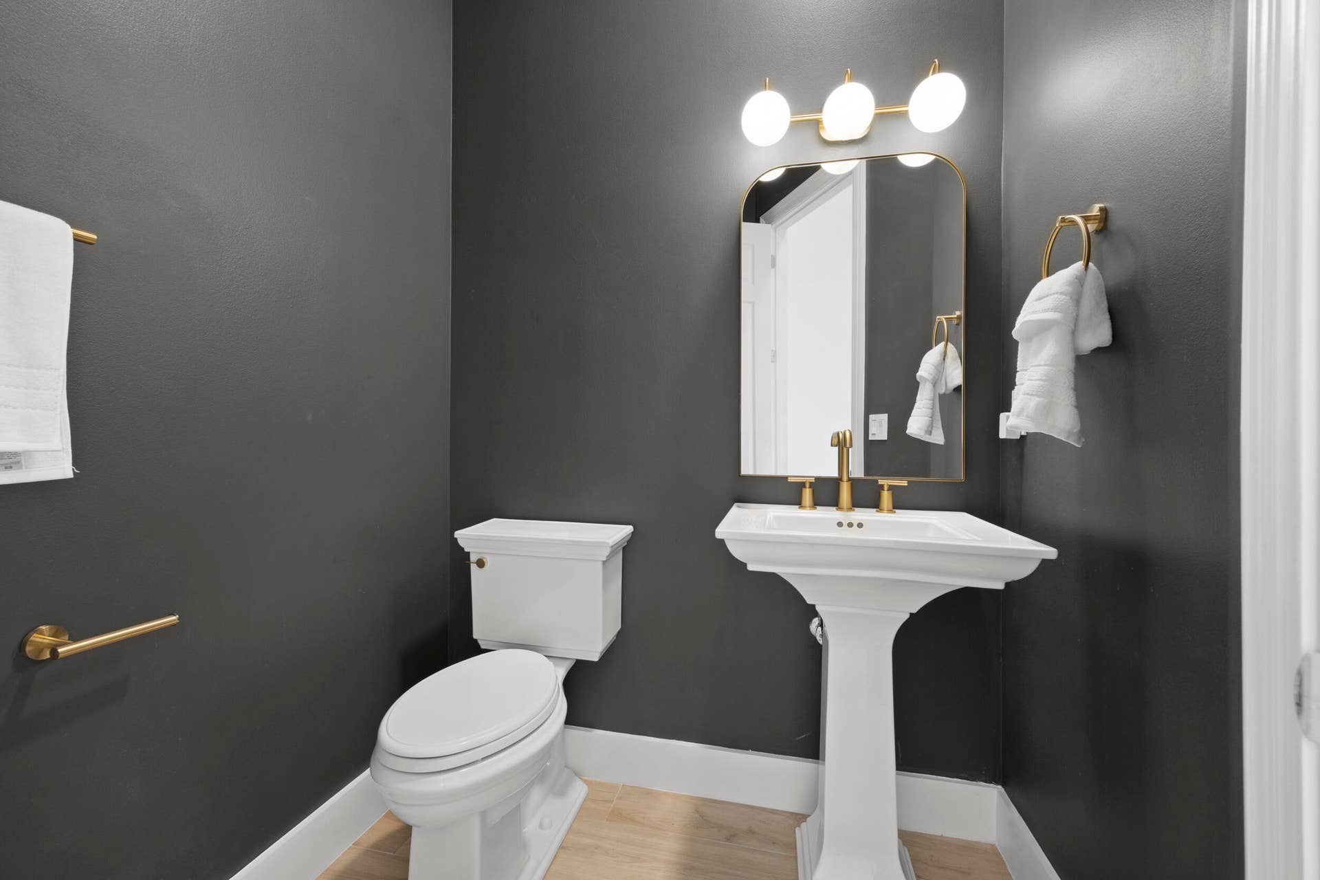 Powder room featuring a white pedestal sink, brass-finish faucet, arched mirror, three-globe light fixture, and wood-finish flooring