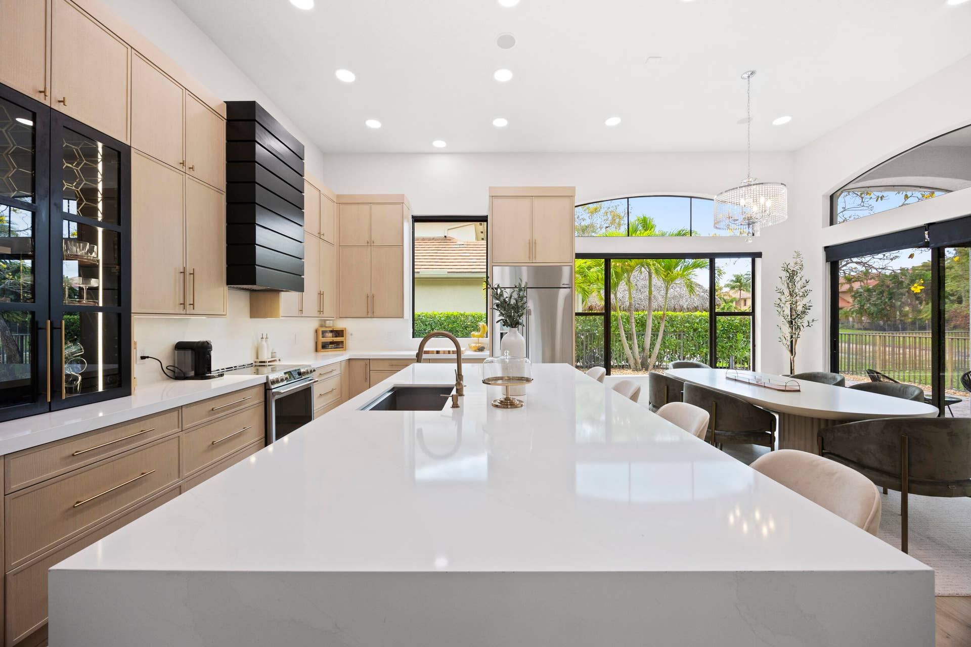 Kitchen featuring light wood-finish cabinetry, a large waterfall-edge quartz island, a high-contrast black range hood, glass-front display cabinetry, and oversized arched windows