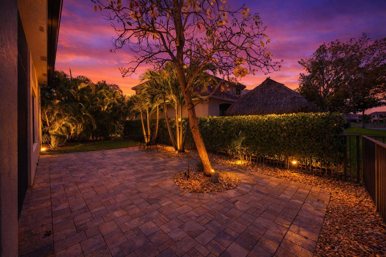 Paver patio featuring a built-in thatched-roof structure, mature landscaping, and integrated landscape lighting
