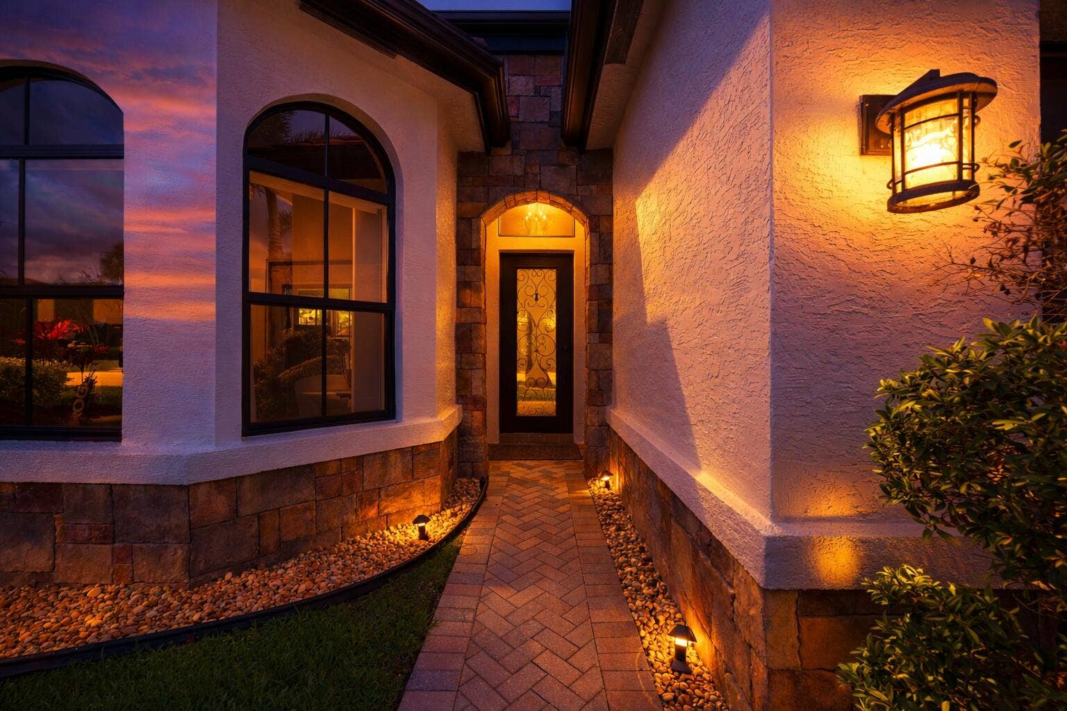 Exterior entry featuring stucco walls with stone veneer accents, arched windows, herringbone-pattern paver walkway, and decorative wall-mounted lanterns