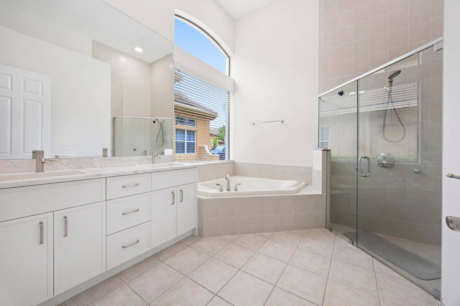 Bathroom featuring a dual-sink vanity with light stone countertops, a large glass-enclosed shower, a deep soaking tub set within an arched window alcove, and tile flooring