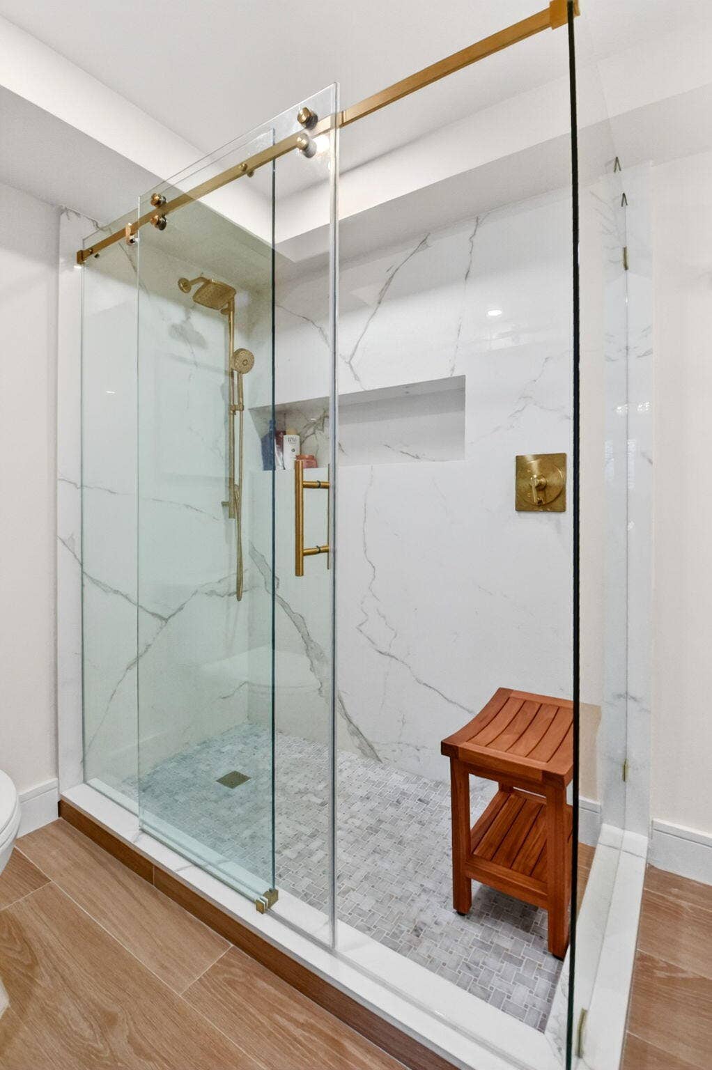 Walk-in shower featuring glass sliding doors, brass-finish hardware, floor-to-ceiling stone-style wall tile, a recessed wall niche, and a pebble-texture tile floor