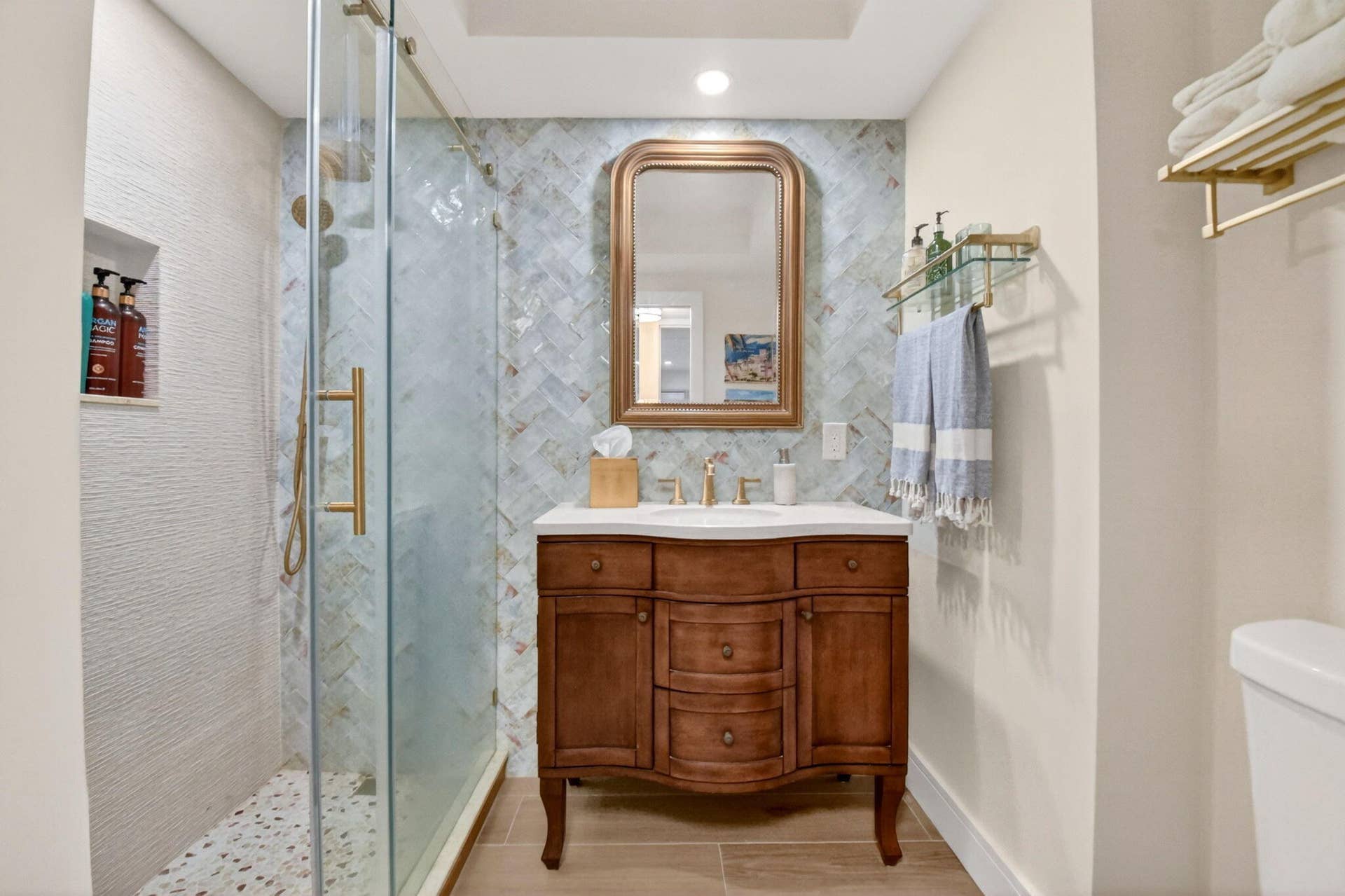 Bathroom featuring a wood vanity with a stone countertop, herringbone-patterned wall tile, glass-enclosed shower with a pebble tile floor, and brass-finish hardware