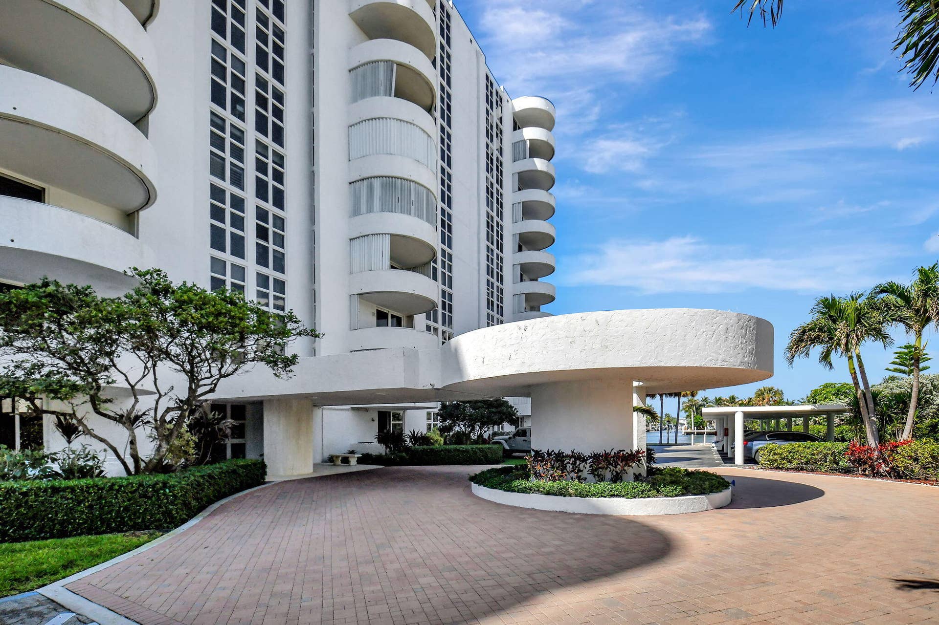 Multi-story residential exterior featuring a circular porte-cochre, curved private balconies, paver stone driveway, and established landscape beds