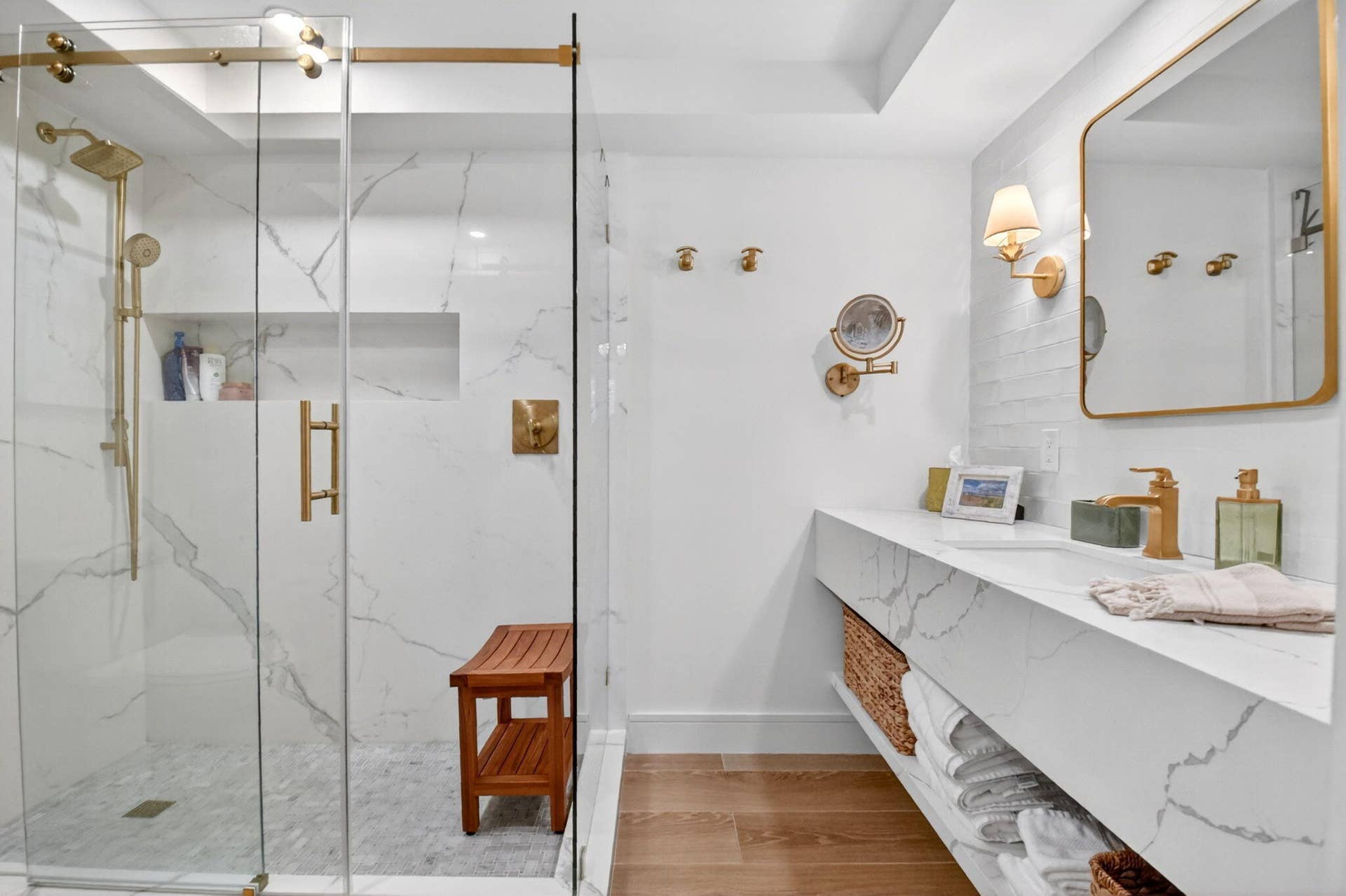 Glass-enclosed walk-in shower with floor-to-ceiling stone-patterned tile, integrated recessed shelving, floating vanity with stone-patterned countertop, gold-toned hardware, and wood-finish flooring