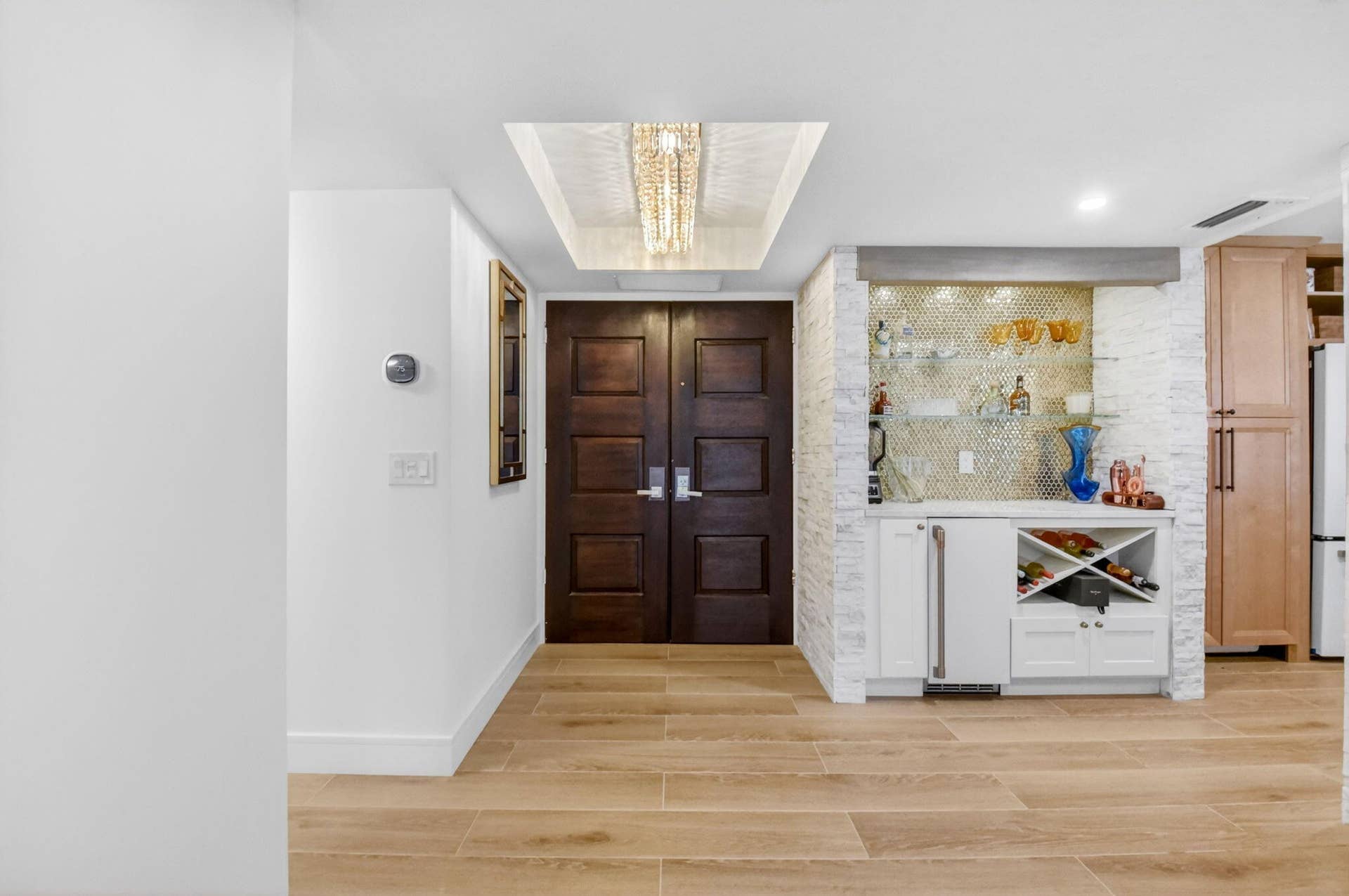 Double door entrance featuring wood-finish flooring, recessed ceiling lighting with crystal chandelier, built-in wet bar with stone-finish backsplash, glass shelving, and under-counter beverage refrigeration