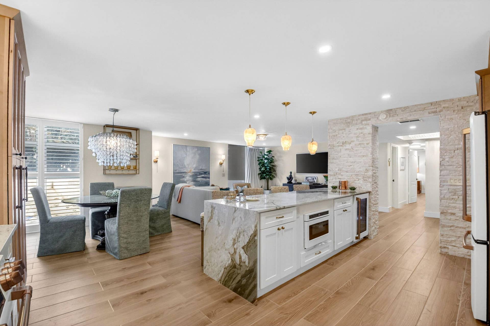 Open-concept kitchen featuring an expansive island with waterfall-edge stone countertops, custom white cabinetry, built-in microwave, integrated wine cooler, and natural wood-finish flooring complemented by a textured stone accent wall