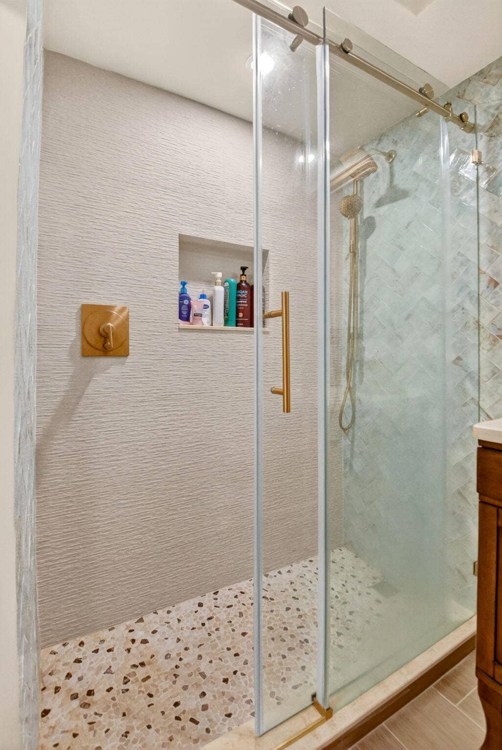 Walk-in shower featuring a frameless sliding glass door, textured wall tile, herringbone-patterned accent tile, a built-in recessed wall niche, and brushed gold finish hardware