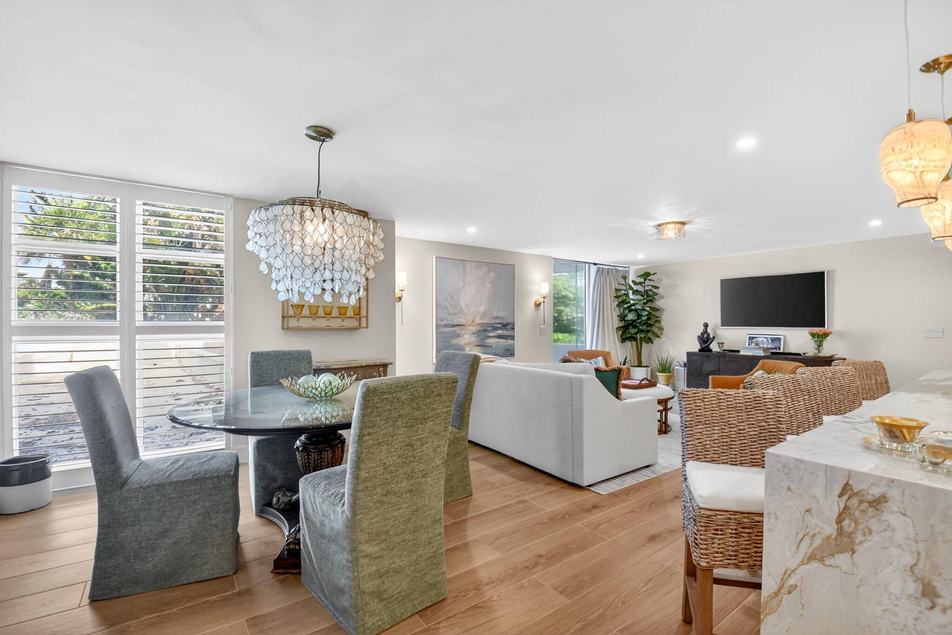 Open-concept living space featuring wood-finish flooring, recessed lighting, ornate multi-tiered chandelier, breakfast bar with stone-finish waterfall countertop, and floor-to-ceiling windows with integrated shutters