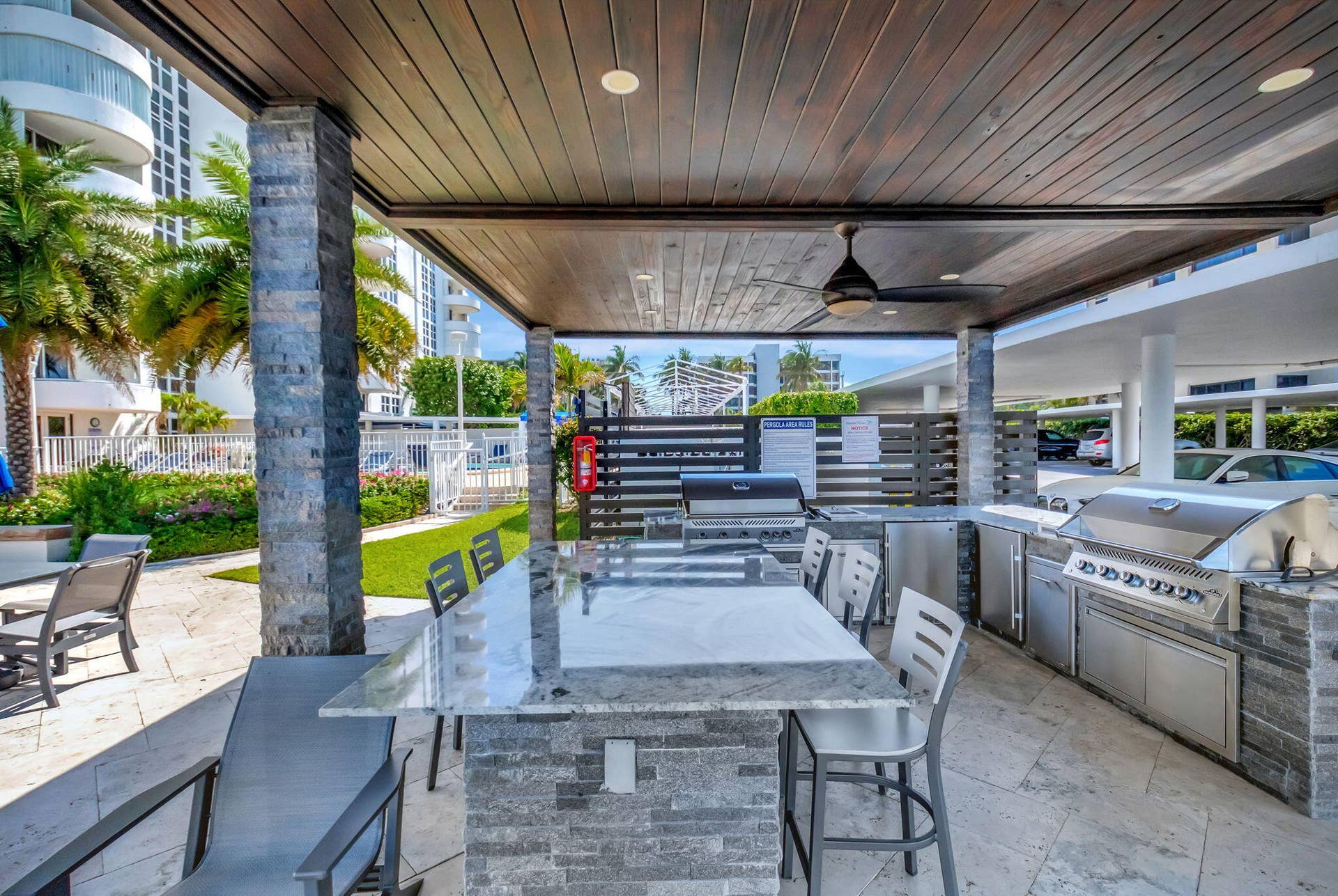 Covered outdoor kitchen featuring stone veneer cabinetry, integrated stainless steel appliances, a stone countertop island, and wood-paneled ceiling with recessed lighting