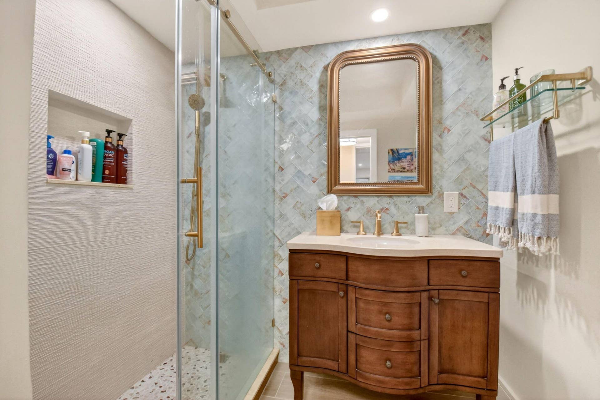 Bathroom vanity with a stone countertop, herringbone-patterned wall tile, glass-enclosed shower with brass fixtures, and a recessed shower niche