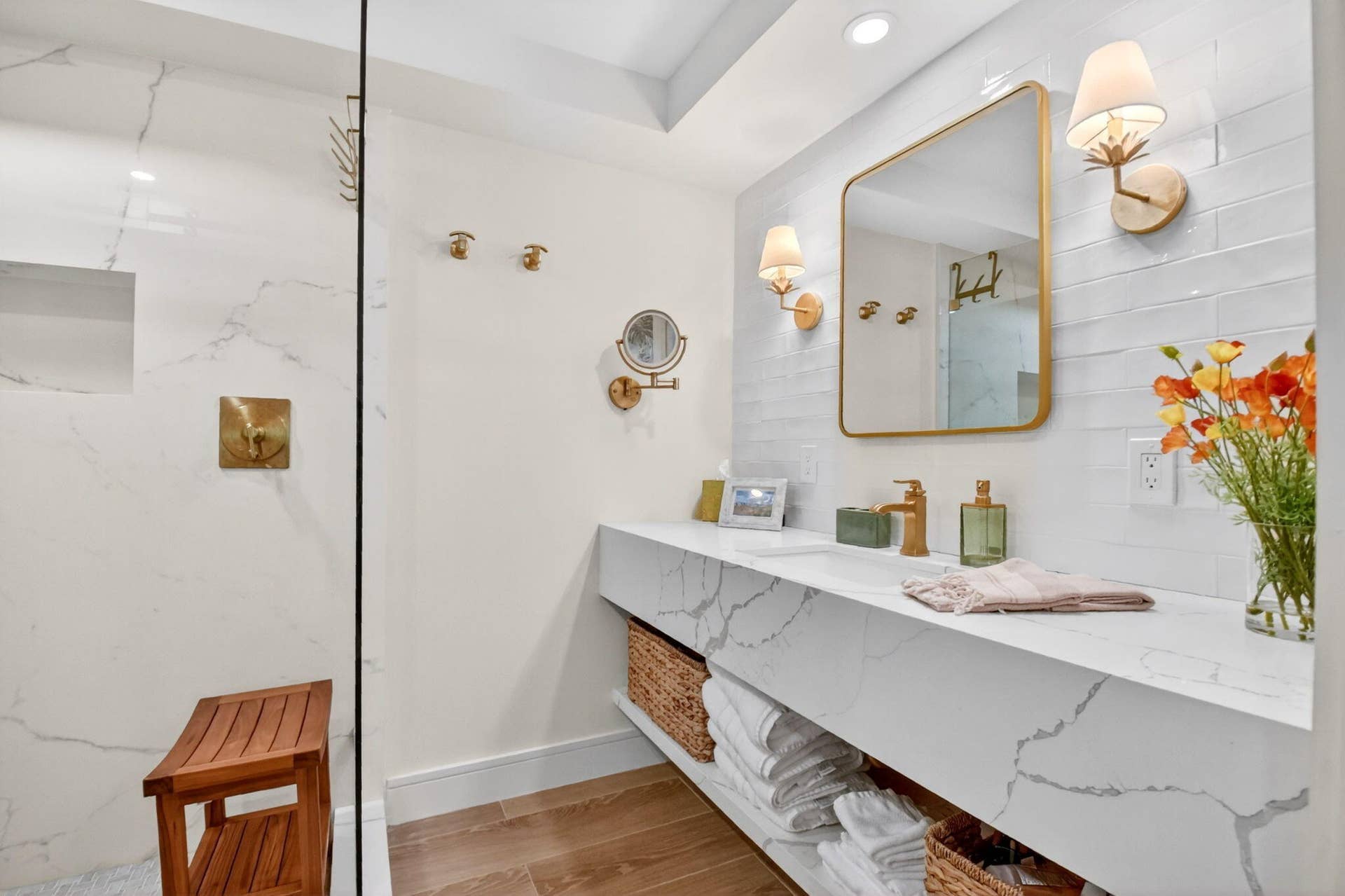 Floating vanity with stone-style countertop, gold-tone fixtures, wall-mounted sconce lighting, frameless glass shower enclosure, and tiled walk-in shower with inset wall niche