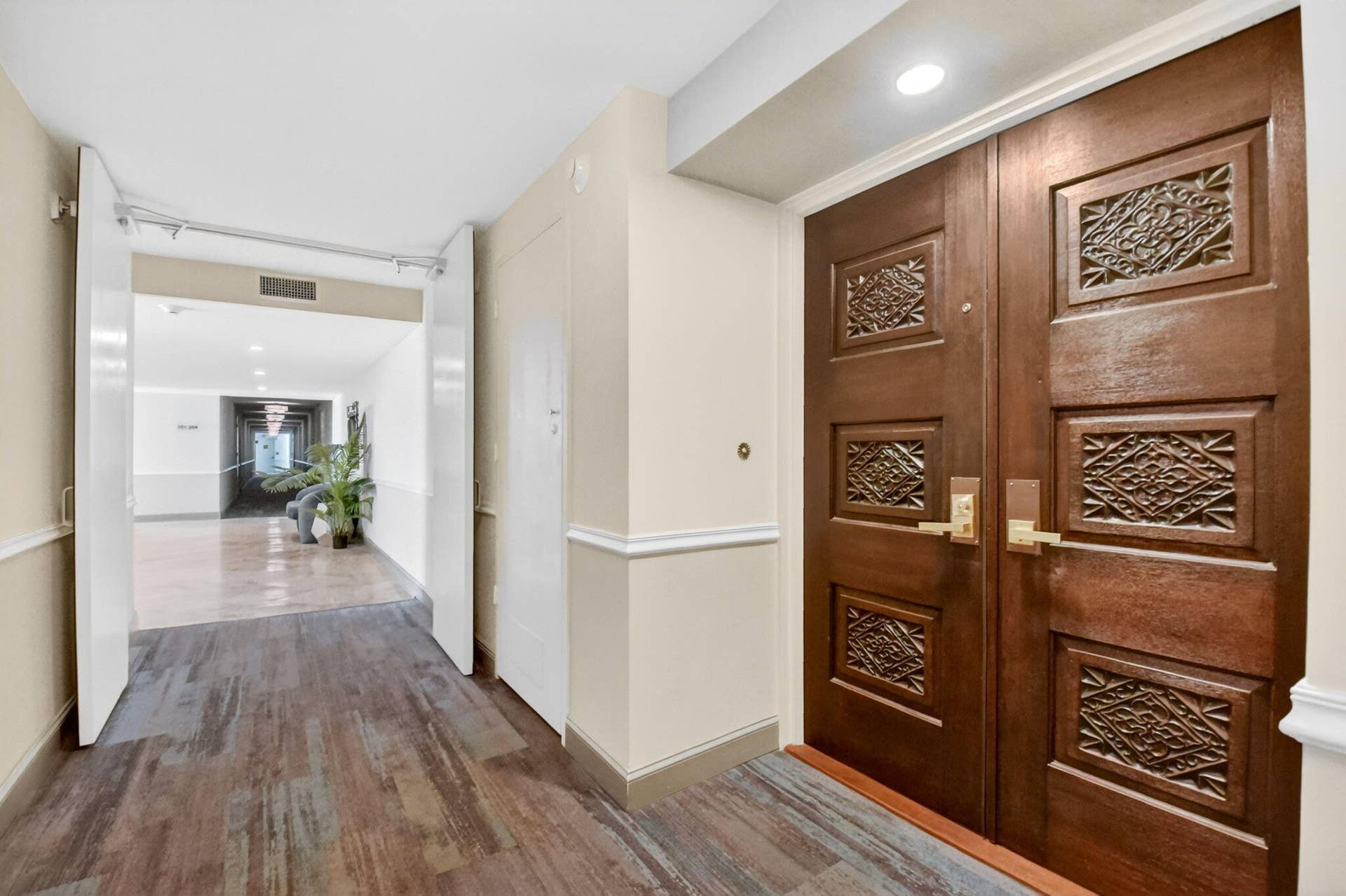 Double entry doors with detailed wood carvings, brass-finish hardware, recessed ceiling lighting, and wood-finish plank flooring