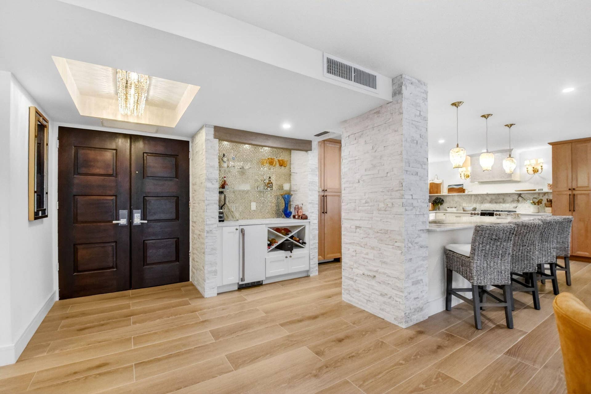 Double-door entrance featuring a recessed ceiling tray with an integrated chandelier, complemented by wood-finish tile flooring throughout, a stacked stone accent pillar, and a built-in wet bar with a wine rack