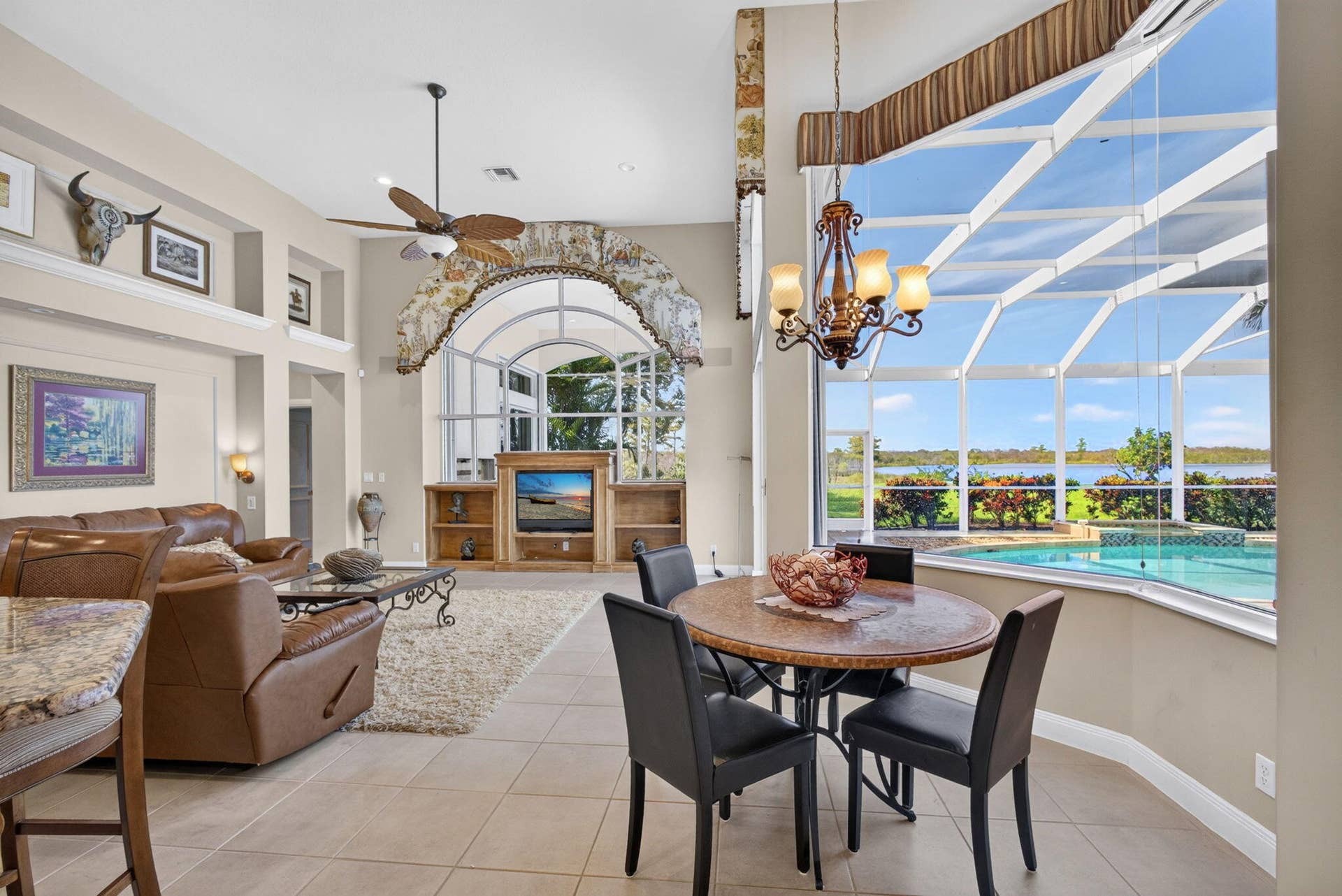42 Windward Isle, Palm Beach Gardens, FL 33418 Open-concept interior featuring large-format tile flooring, a custom arched built-in media center, high ceilings with a ceiling fan, and a sprawling window wall enclosure showcasing water frontage and an in-ground pool