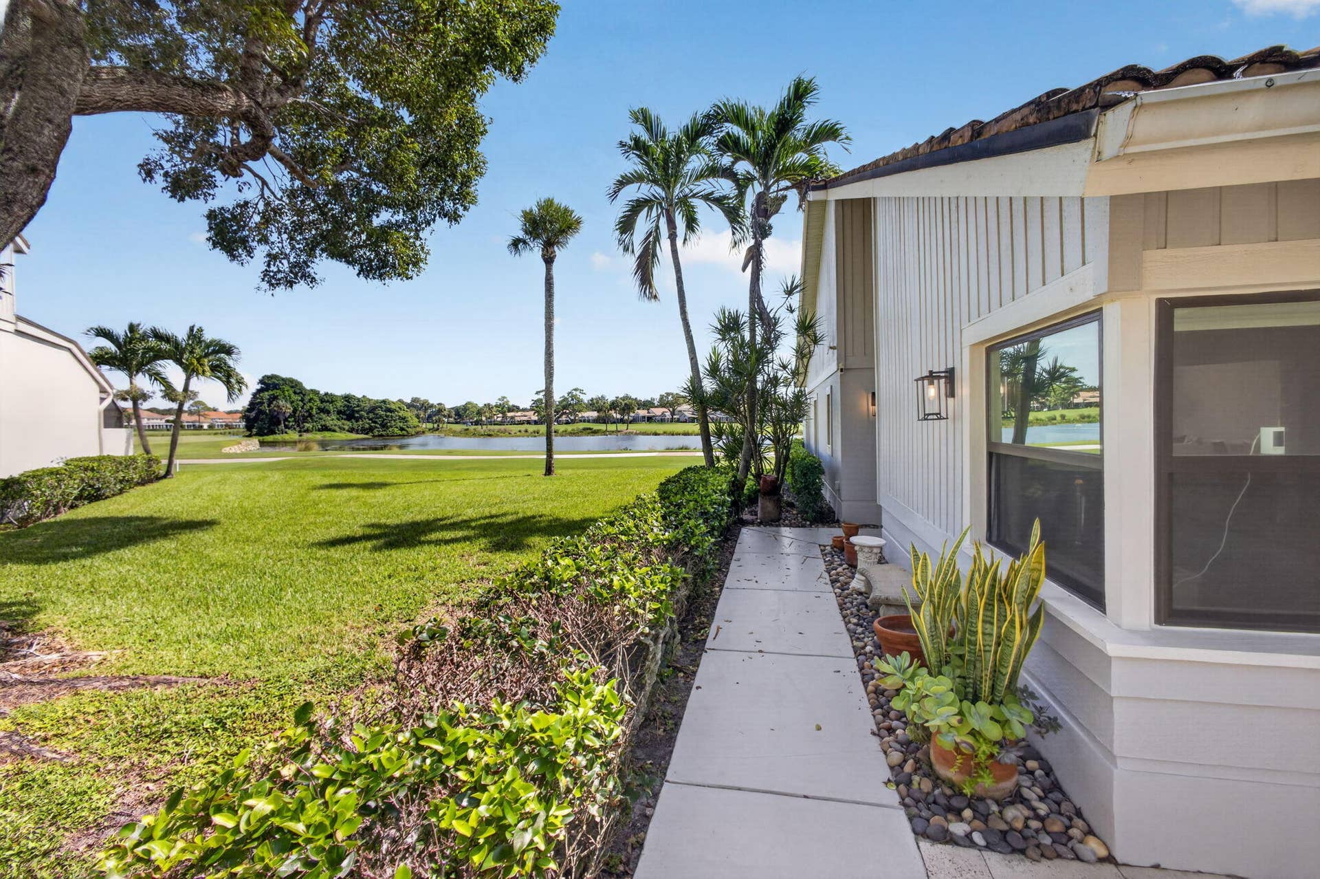 543 Prestwick Circle, Palm Beach Gardens, FL 33418 Exterior concrete walkway bordered by manicured hedging and stone ground cover, featuring vertical siding, metal wall-mounted lighting, and grounds overlooking a pond
