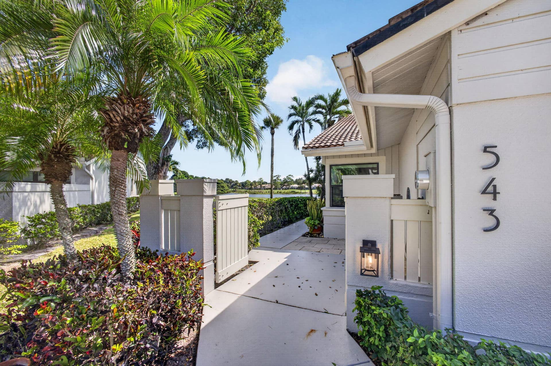 543 Prestwick Circle, Palm Beach Gardens, FL 33418 Exterior entrance featuring a concrete walkway, tiled roof, wall-mounted lantern-style lighting, and mature palm landscaping