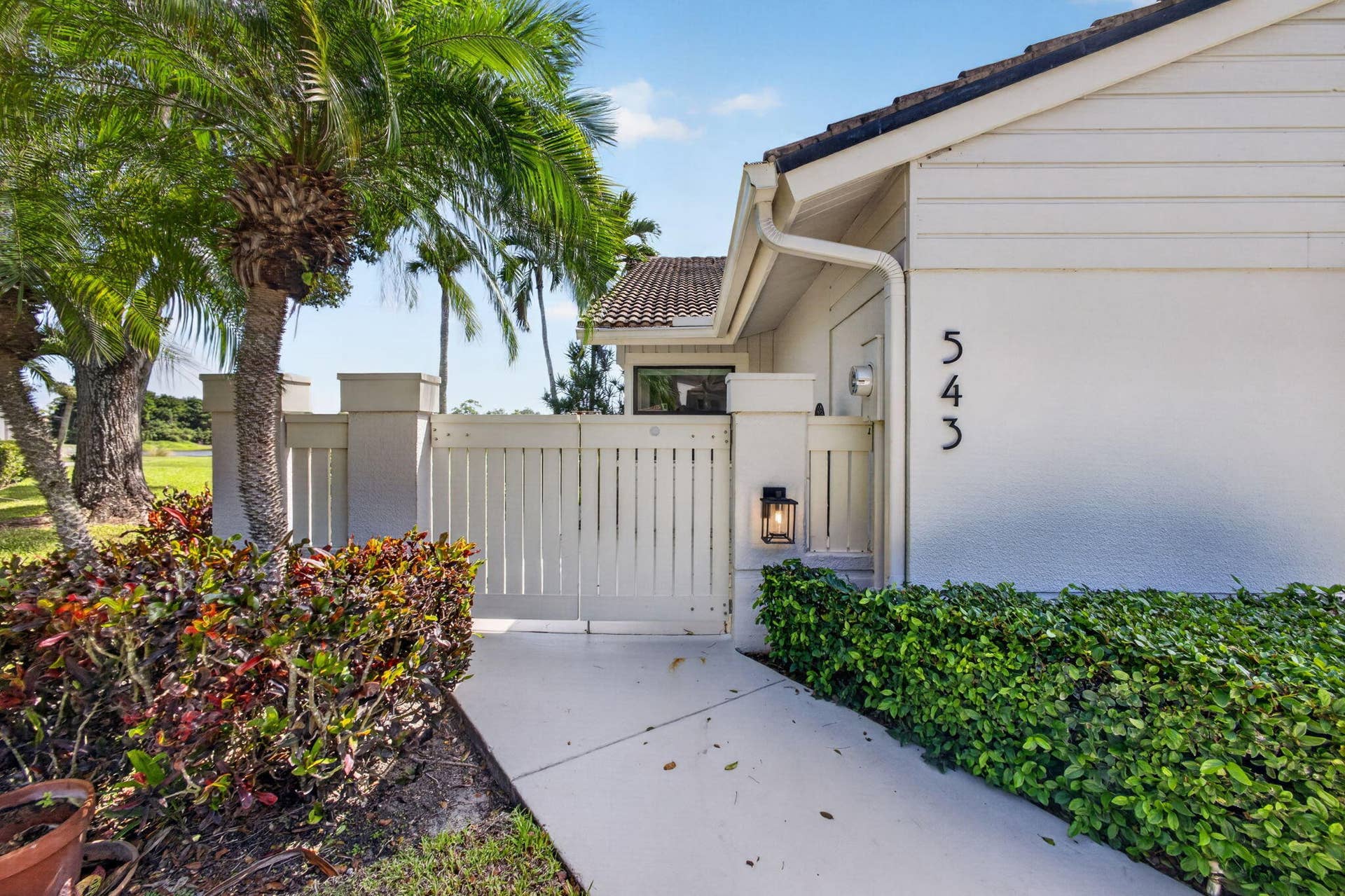 543 Prestwick Circle, Palm Beach Gardens, FL 33418 Exterior entrance featuring a concrete walkway, gated courtyard with privacy fencing, integrated wall-mounted lighting, and mature palm tree landscaping