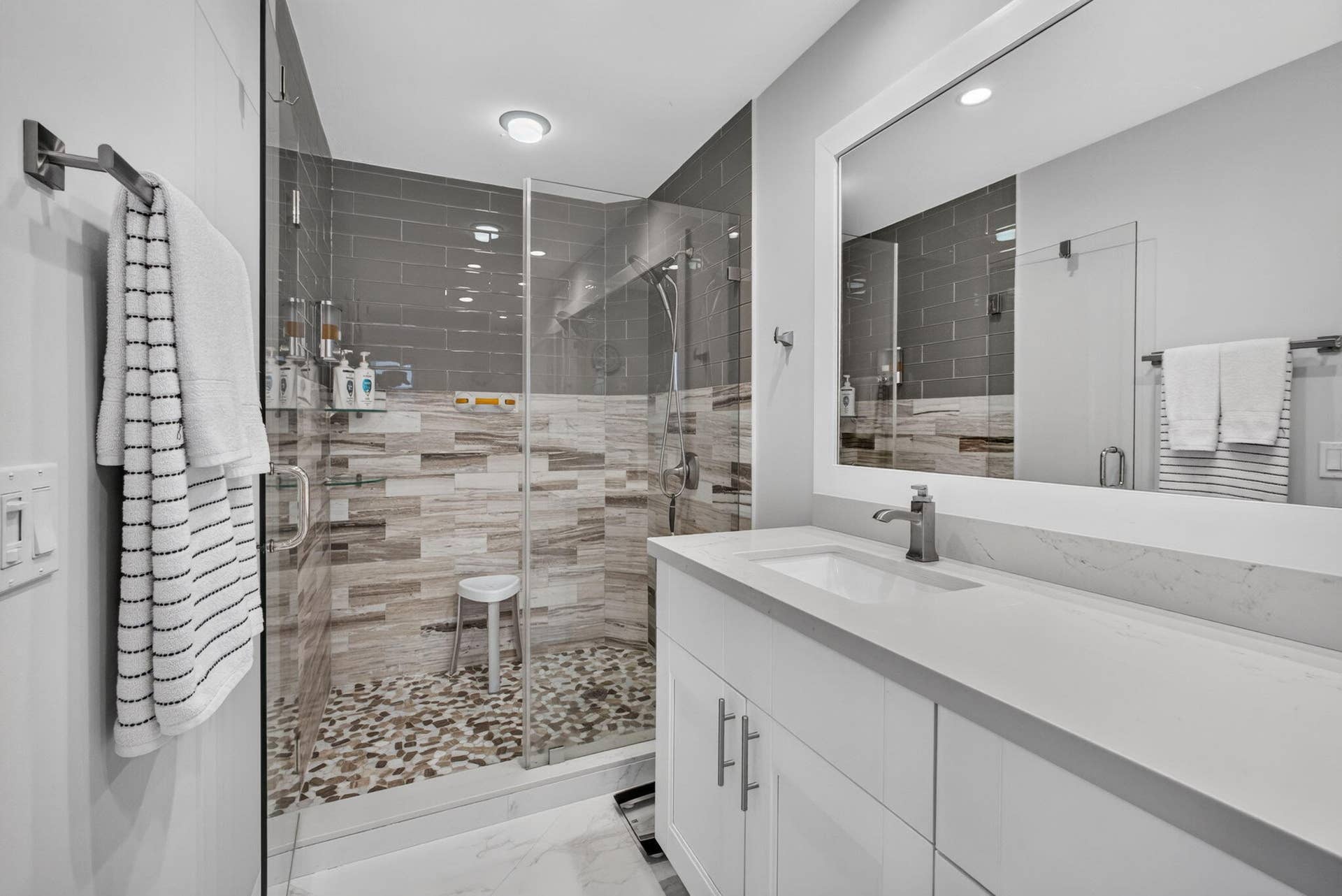 543 Prestwick Circle, Palm Beach Gardens, FL 33418 Bathroom featuring a white vanity with a light-toned countertop, a walk-in shower with a glass enclosure, decorative multi-toned tile work, and a pebble-stone shower floor
