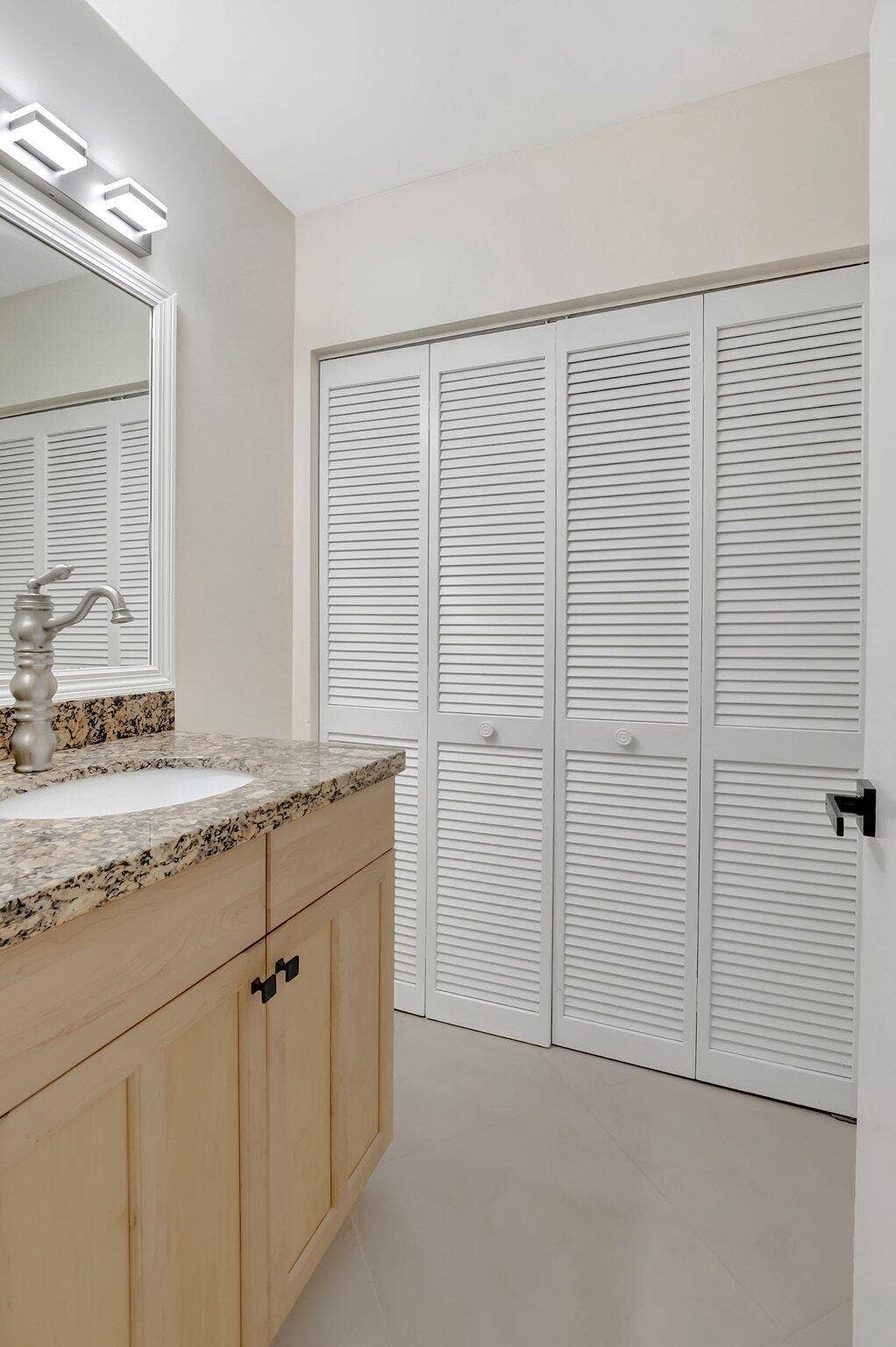 Bathroom vanity featuring a light-toned wood cabinet, stone-finish countertop with an integrated basin, high-arc faucet, wall-mounted light fixture, and floor-to-ceiling louvered closet doors