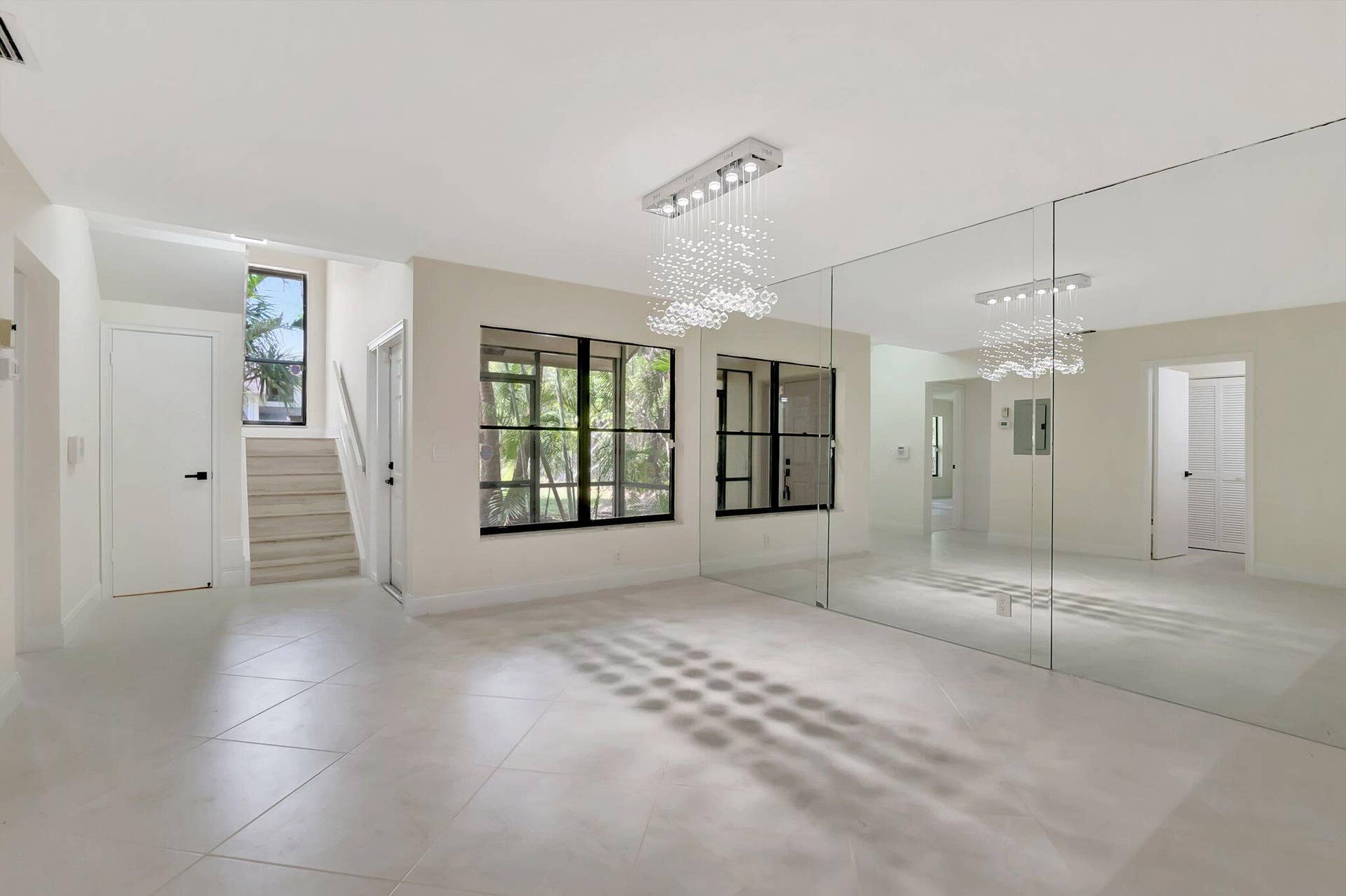 Open-concept interior featuring large-format tile flooring, a floor-to-ceiling mirrored wall, a rectangular multi-light crystal chandelier, black-framed windows, and a set of stairs with carpeted treads