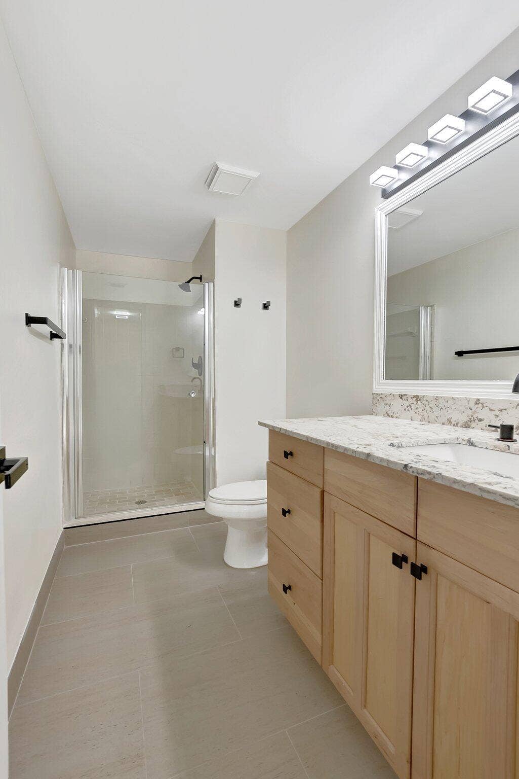 Light-wood cabinetry featuring matte black hardware, stone-finish countertop, walk-in shower with glass enclosure, and large-format tile flooring