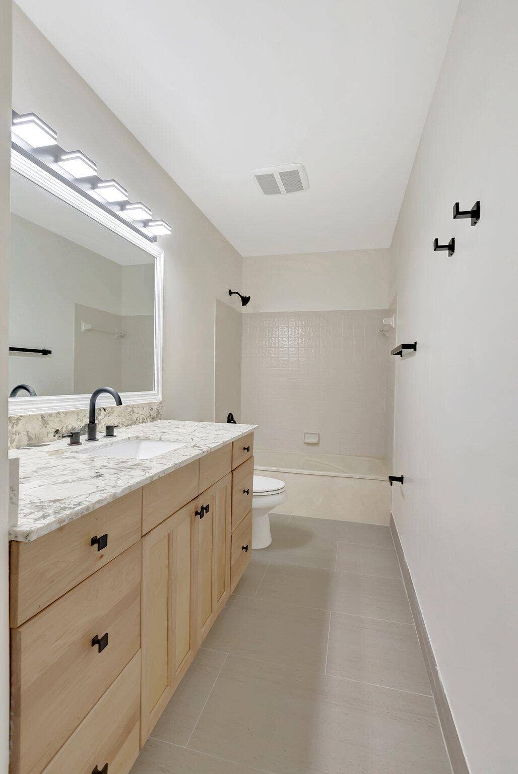 Bathroom featuring a light-finish wood vanity, patterned stone-surface countertop, rectangular undermount sink, matte black hardware, and large-format tile flooring