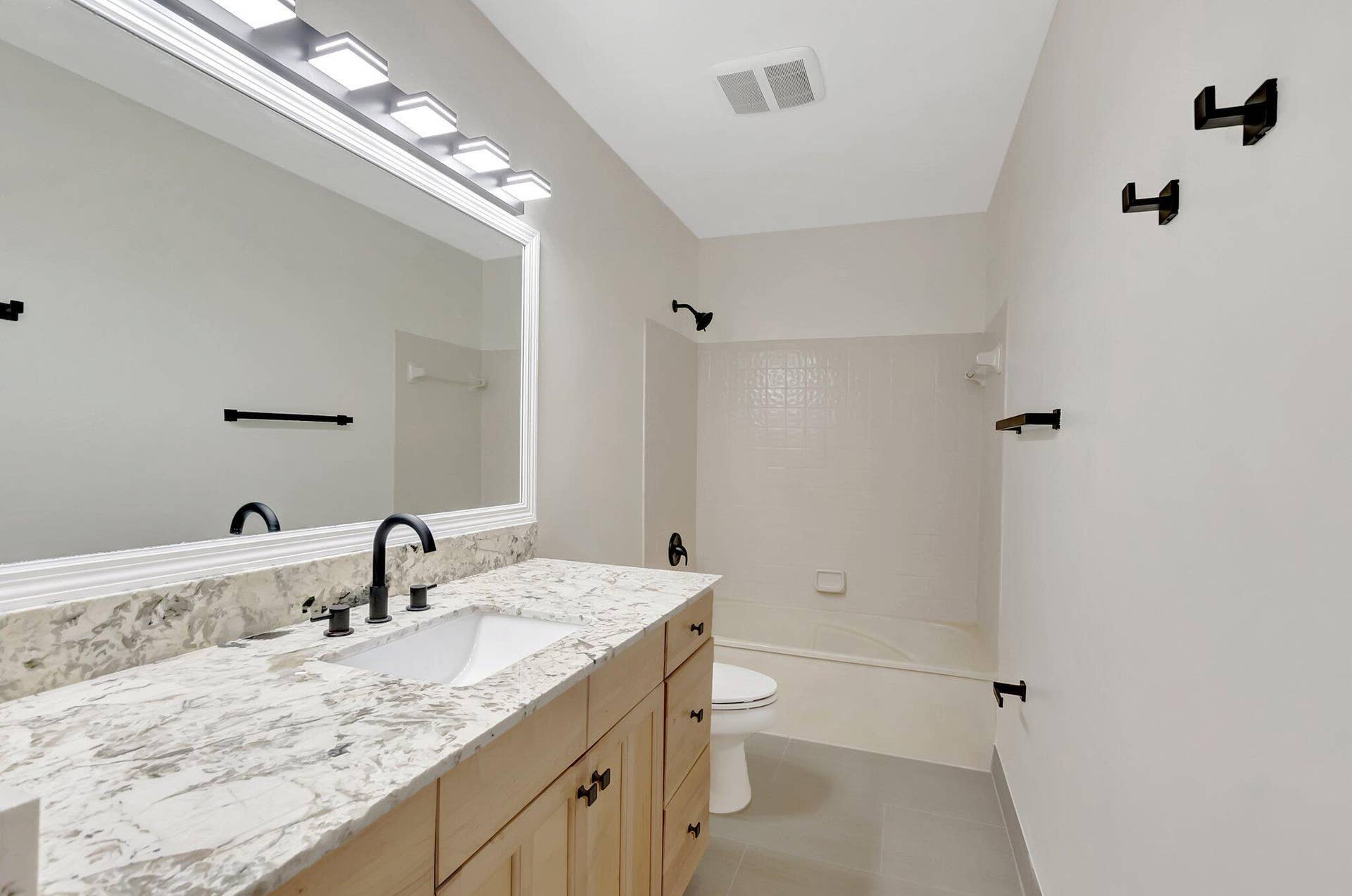 Light-tone wood cabinetry topped with a stone-finish countertop, undermount sink, matte black widespread faucet, expansive wall-to-wall mirror with integrated lighting, and a bathtub-shower combination with matching matte black fixtures
