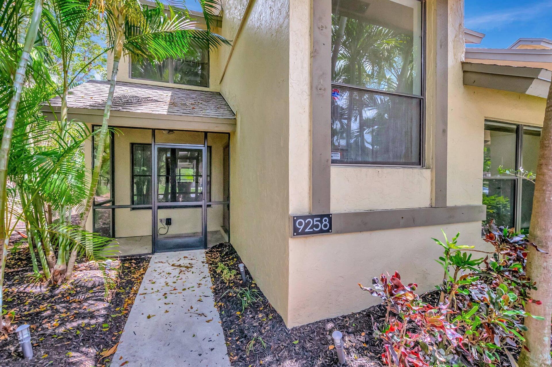 Exterior facade featuring a stucco finish, screened-in entryway, concrete walkway, and mature landscaping