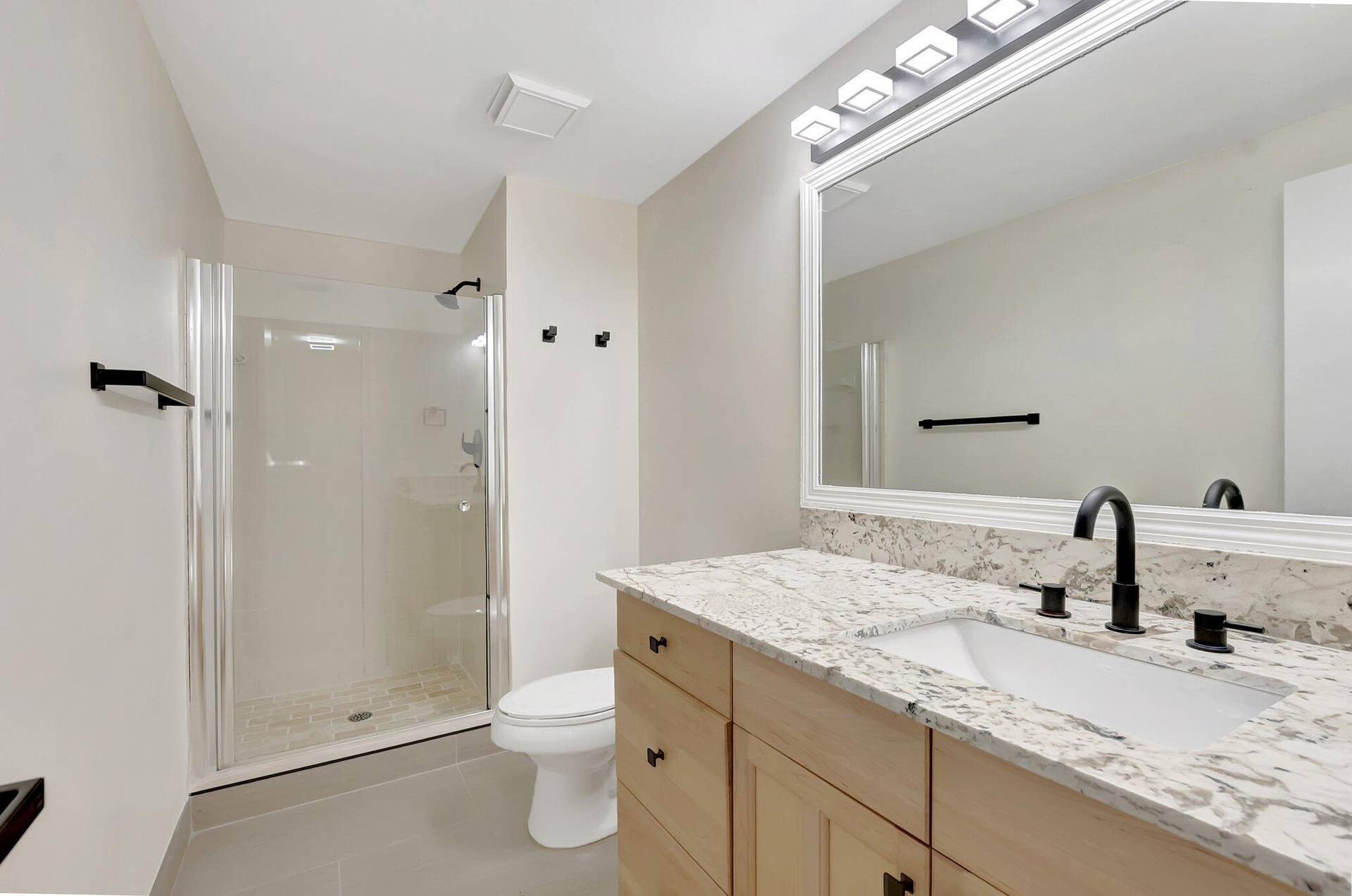 Bathroom featuring a vanity with light wood-finish cabinetry, patterned stone countertop, undermount sink, and matte black hardware, paired with a glass-enclosed shower